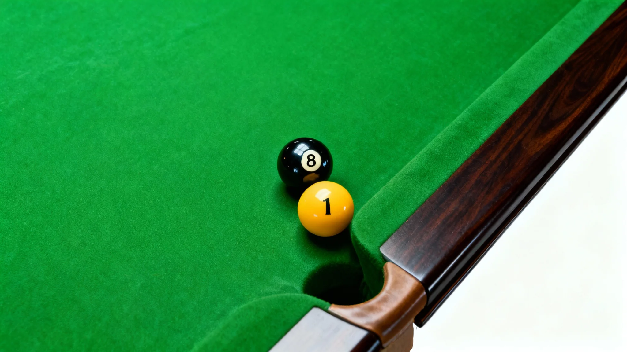 A clean, minimalist image of a pool table corner from a high angle, showing the vibrant green felt, a black 8-ball, a yellow 1-ball, and the polished wood rail against a white background.