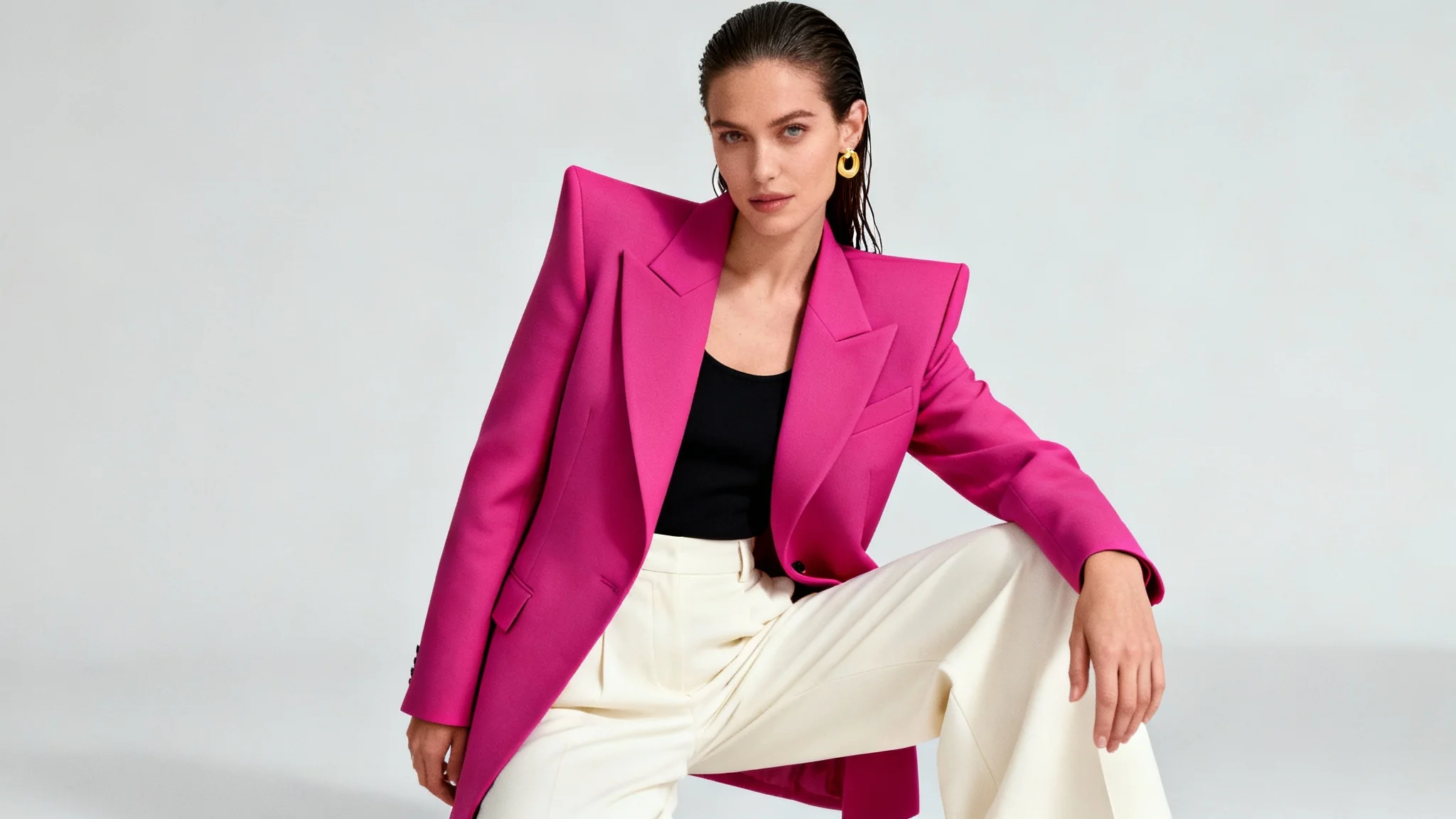 A fashion model poses confidently in a studio, wearing a bright pink blazer, cream trousers, and gold jewelry against a plain light grey background.