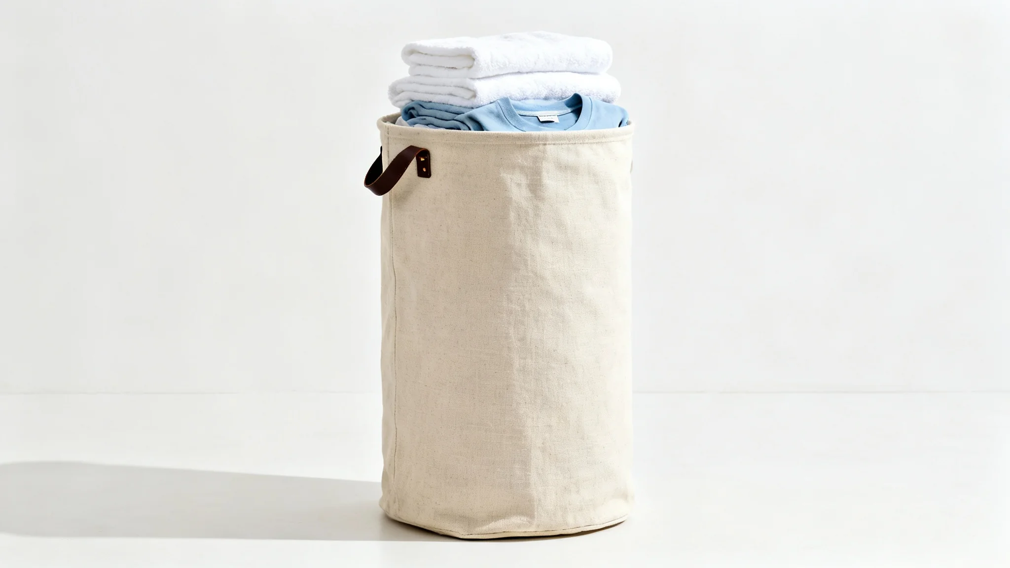 A high-quality product mockup of a beige canvas laundry bag with a leather handle, standing upright on a clean white background, partially filled with neatly folded clean clothes.