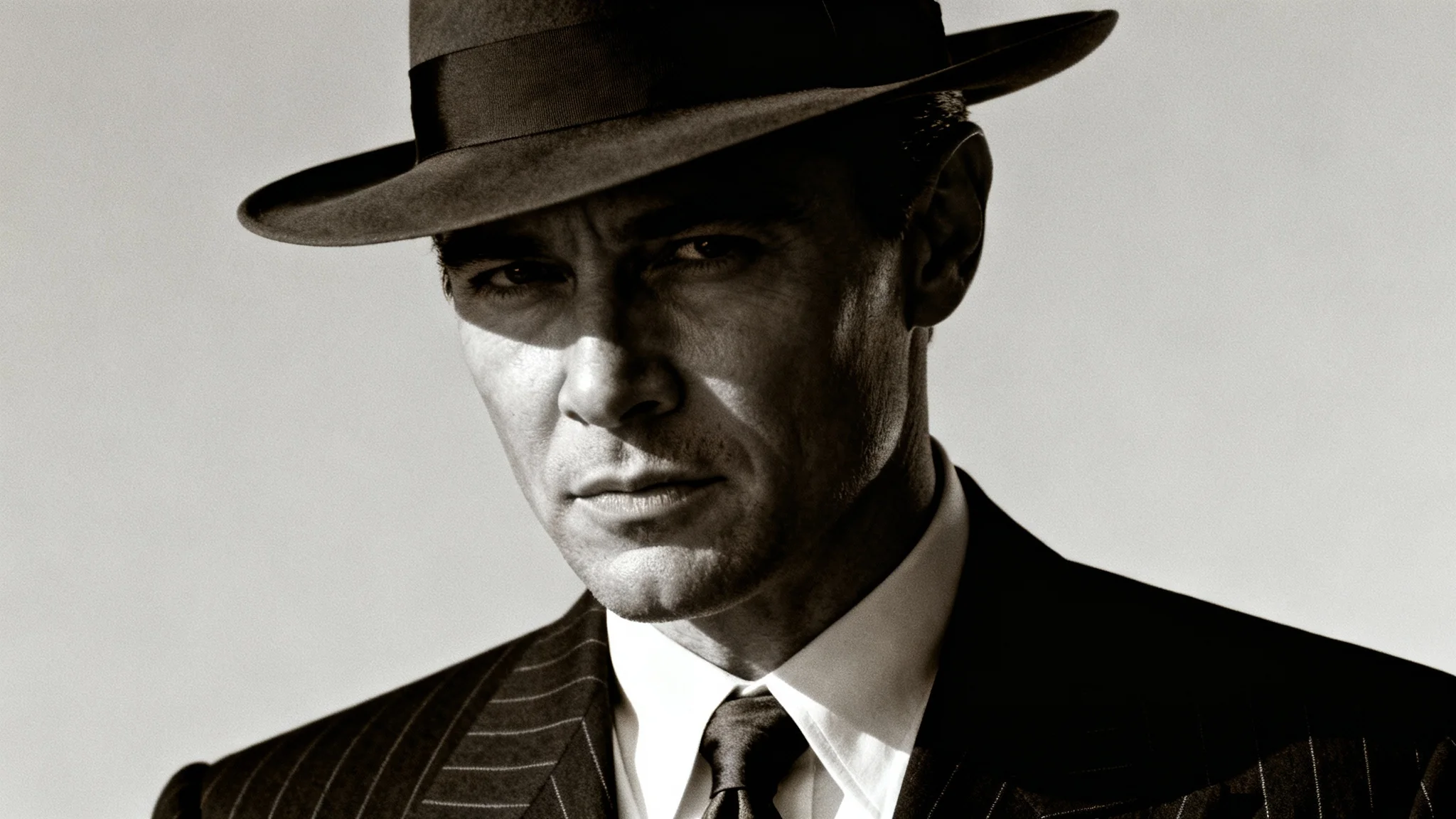 A man dressed as a classic 1940s mobster in a pinstripe suit and fedora, edited in a high-contrast, desaturated film noir style against a plain background.