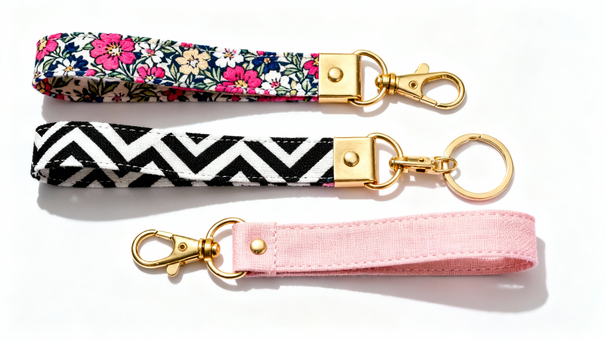 A flat lay photograph of three handmade wristlet key fobs with floral, geometric, and pink linen patterns, all with gold hardware, on a white background.