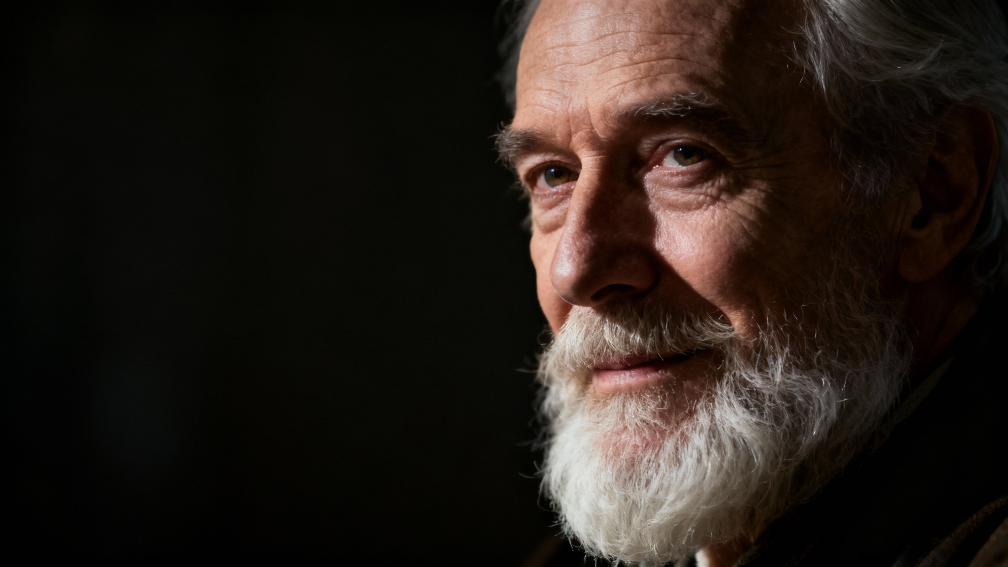 A dramatic, cinematic portrait of an elderly man with a white beard. He is lit with strong side lighting against a dark, blurry background, creating a moody and thoughtful atmosphere.