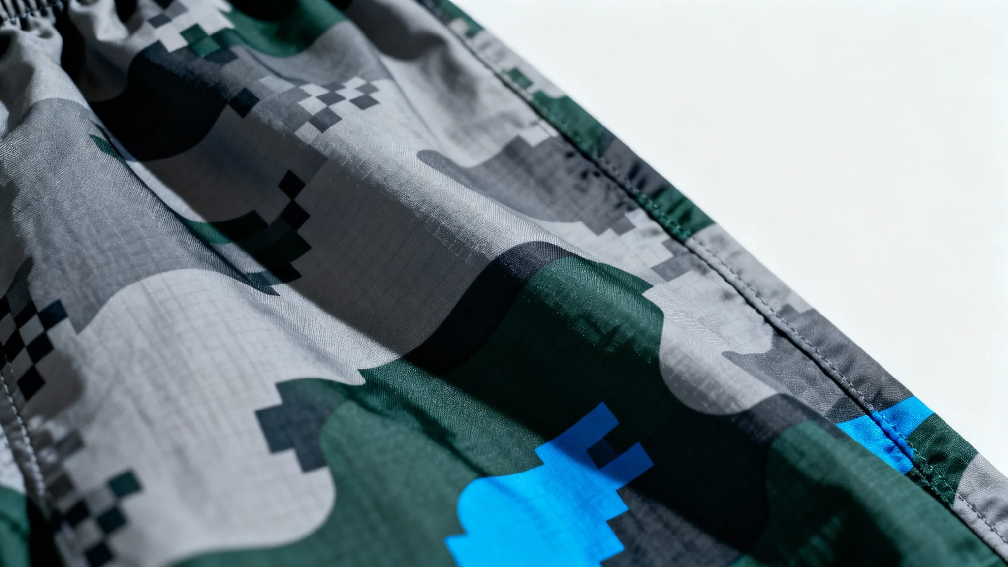 A detailed close-up of a modern camouflage pattern fabric, featuring a mix of digital and organic shapes in gray, green, and blue, set against a plain white background.
