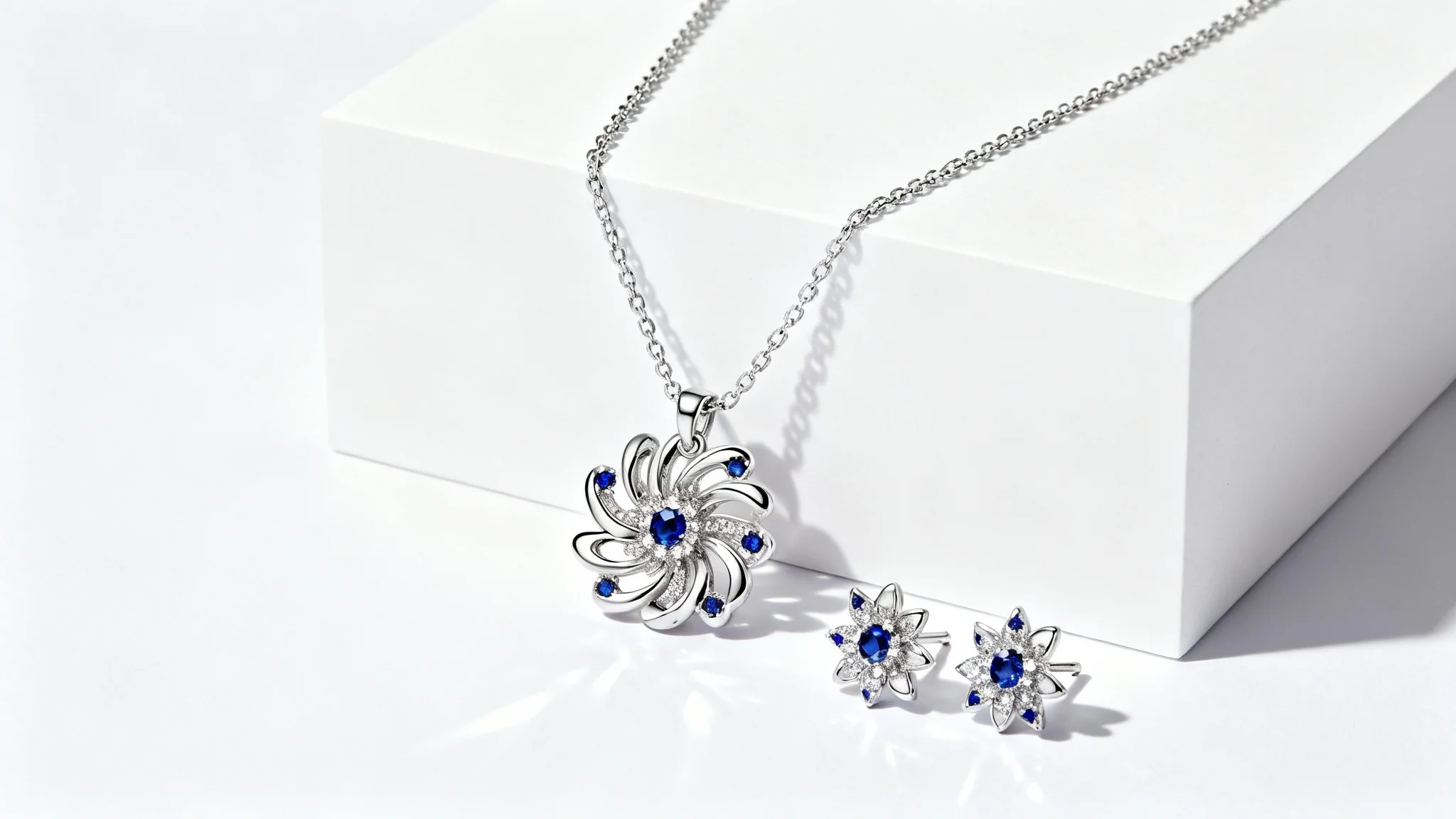 A close-up photograph of a modern, custom-designed silver jewelry set with sapphire details, including a necklace and earrings, displayed elegantly on a white background.