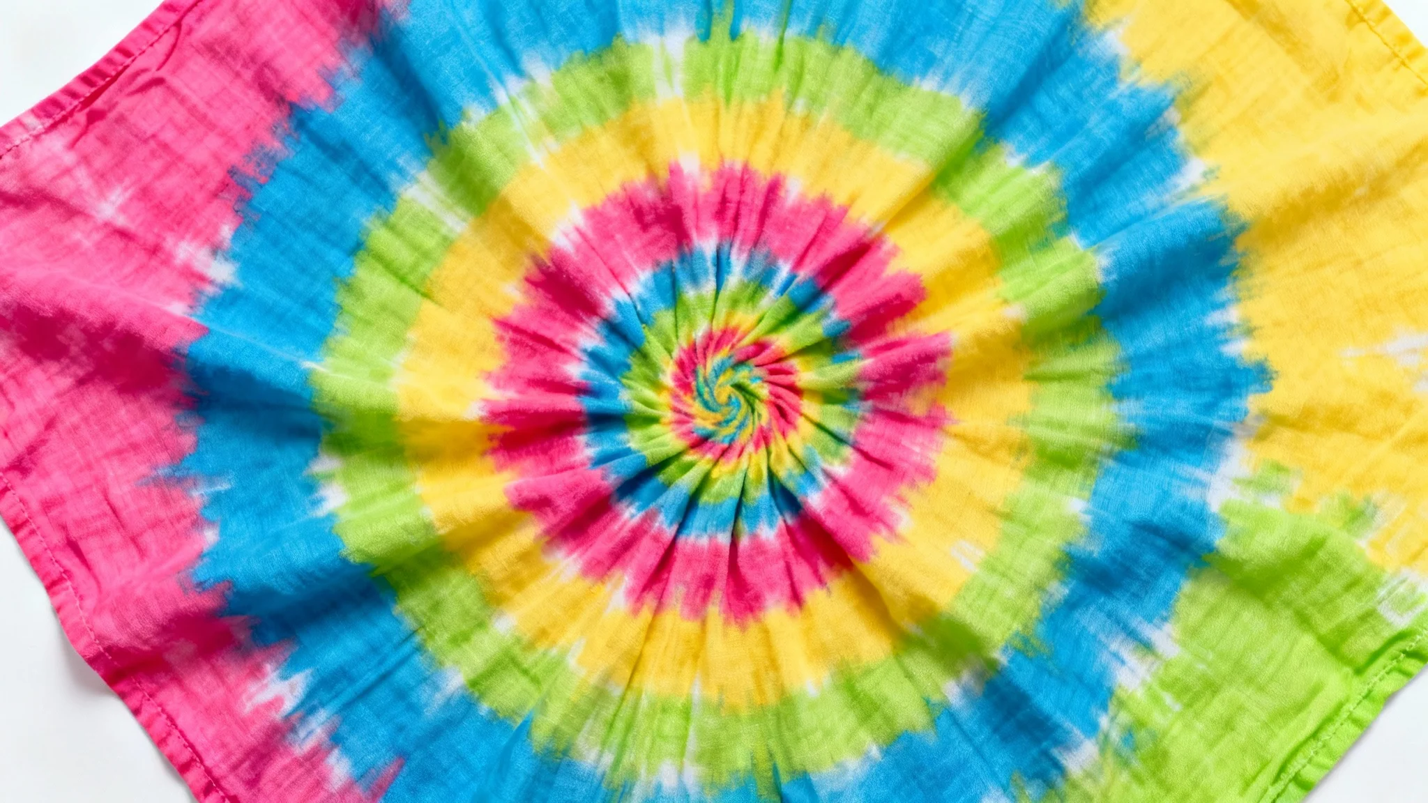 A close-up, top-down view of a vibrant spiral tie-dye pattern on a piece of fabric, showcasing bright colors against a plain white background.