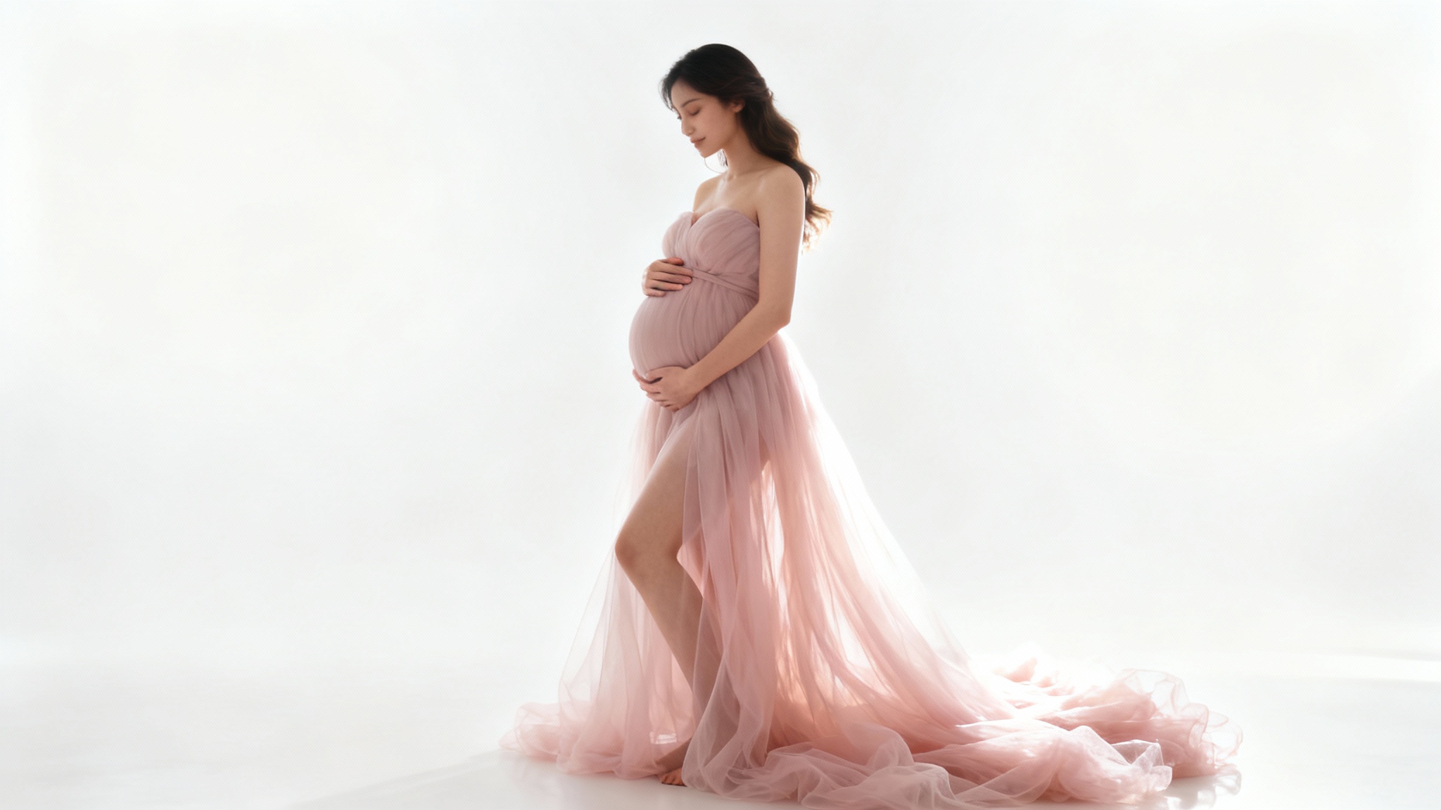 A beautiful pregnant woman posing sideways in a flowing pink gown against a plain white background, gently holding her baby bump during a maternity photoshoot.