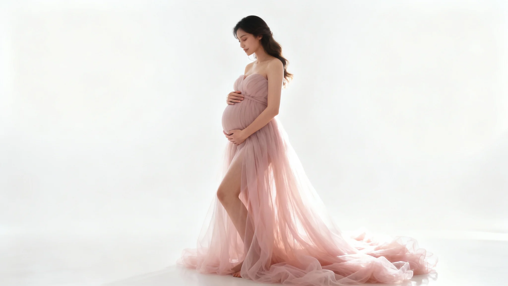 A beautiful pregnant woman posing sideways in a flowing pink gown against a plain white background, gently holding her baby bump during a maternity photoshoot.