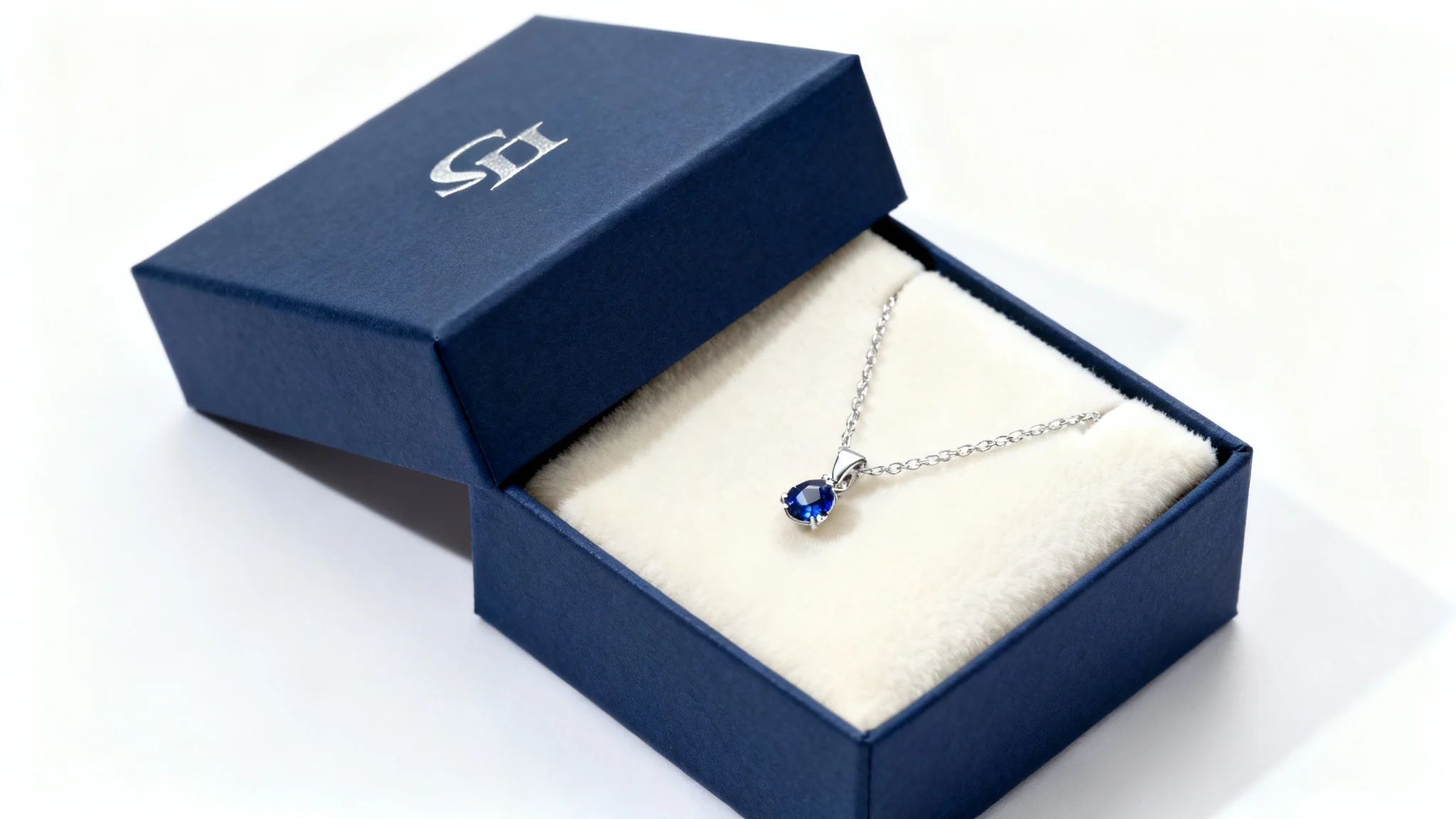 A high-end navy blue jewelry gift box mockup, slightly open to show a silver necklace with a sapphire pendant inside, presented against a clean white background in a studio setting.