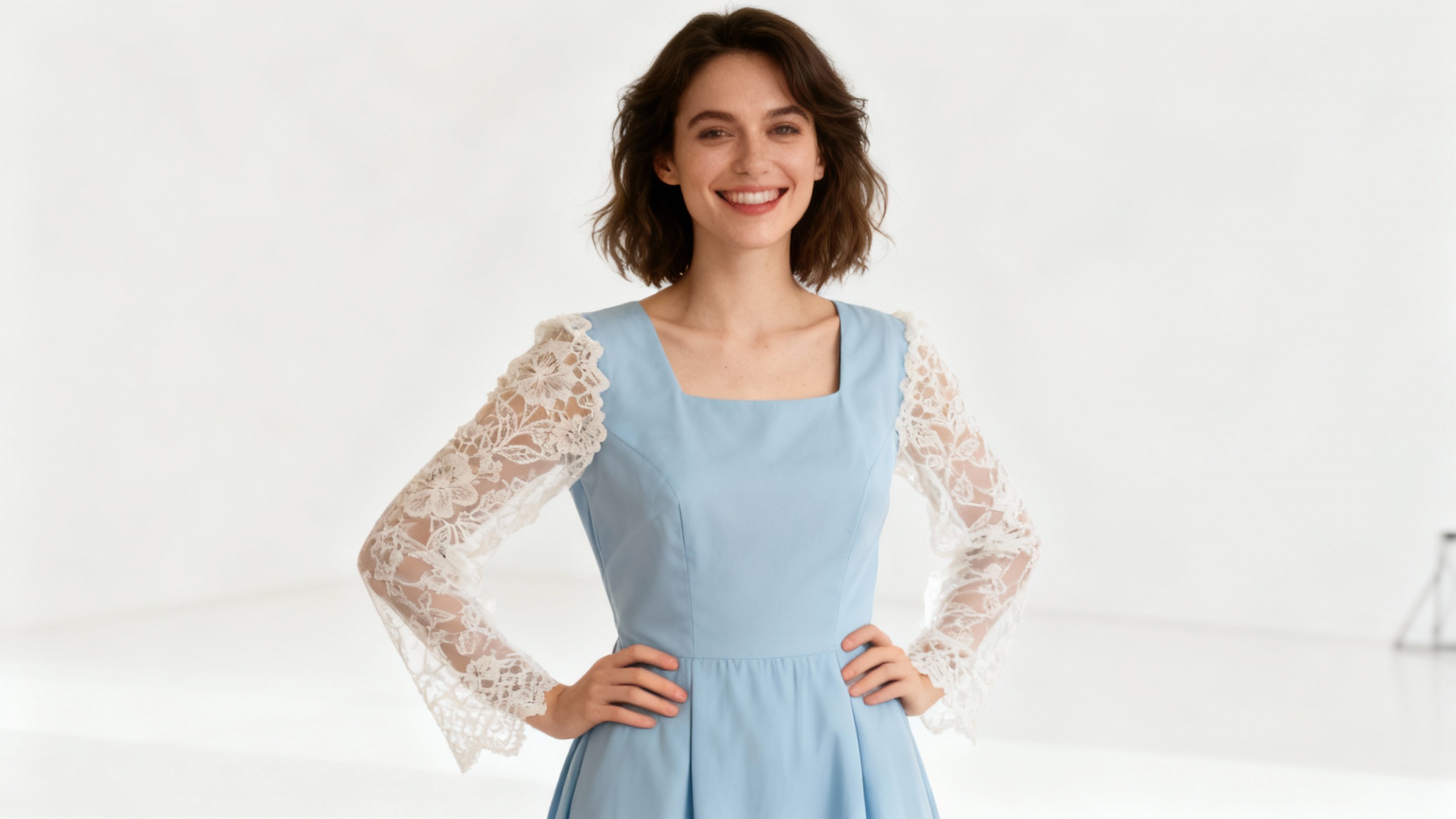 A woman models a light blue dress with elegant white lace long sleeves added to it, standing in a professional photo studio.
