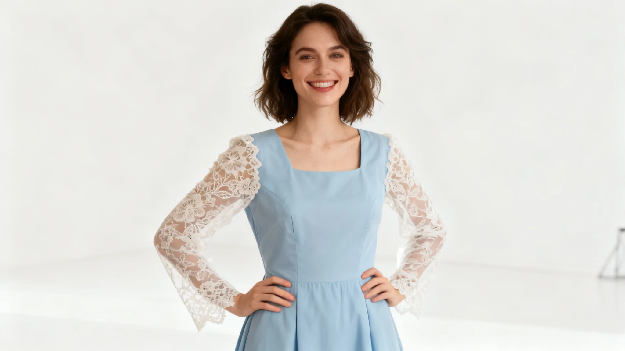 A woman models a light blue dress with elegant white lace long sleeves added to it, standing in a professional photo studio.