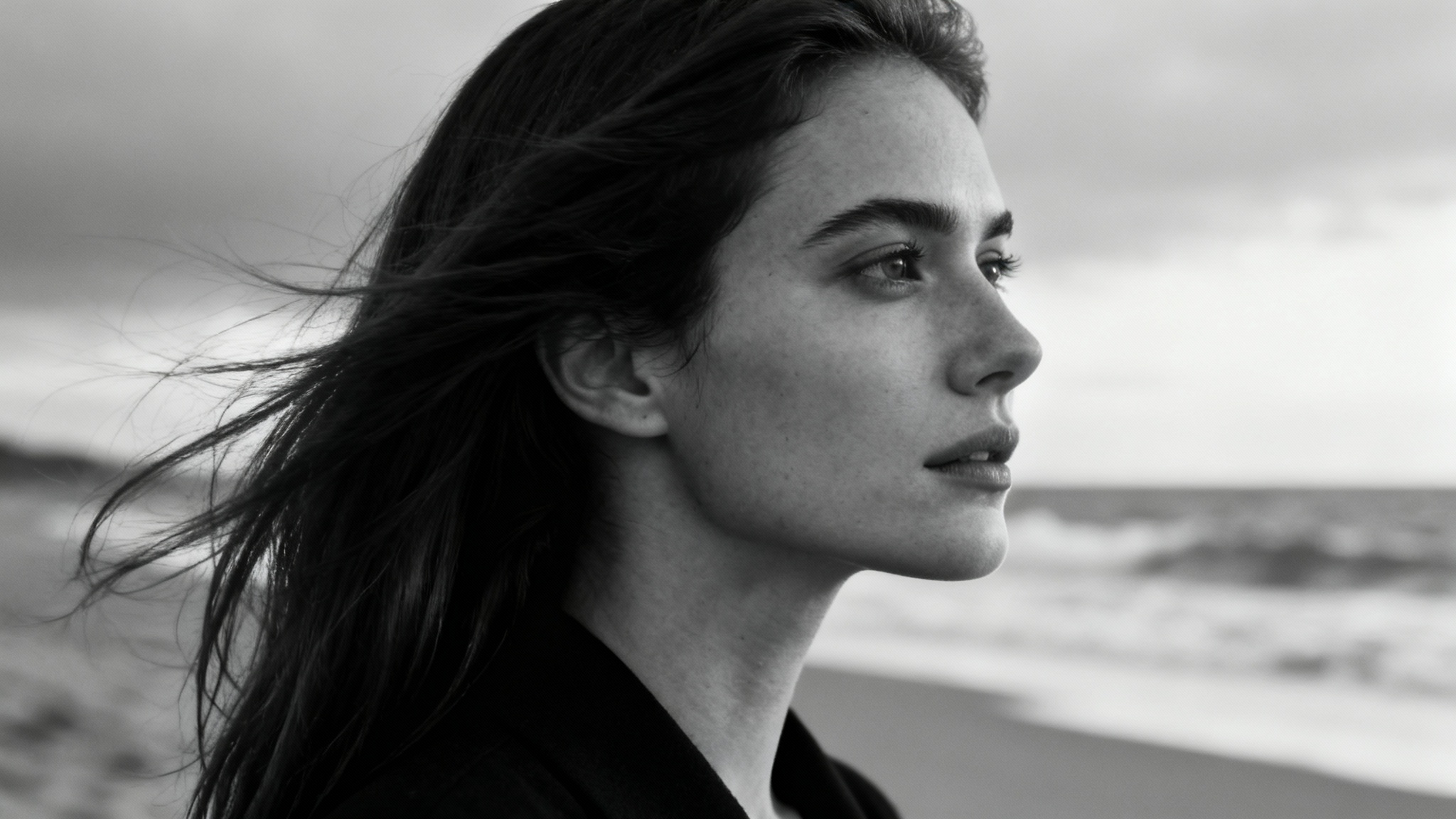 A close-up black and white portrait of a woman on a beach, her hair blowing in the wind as she looks serenely towards the ocean.
