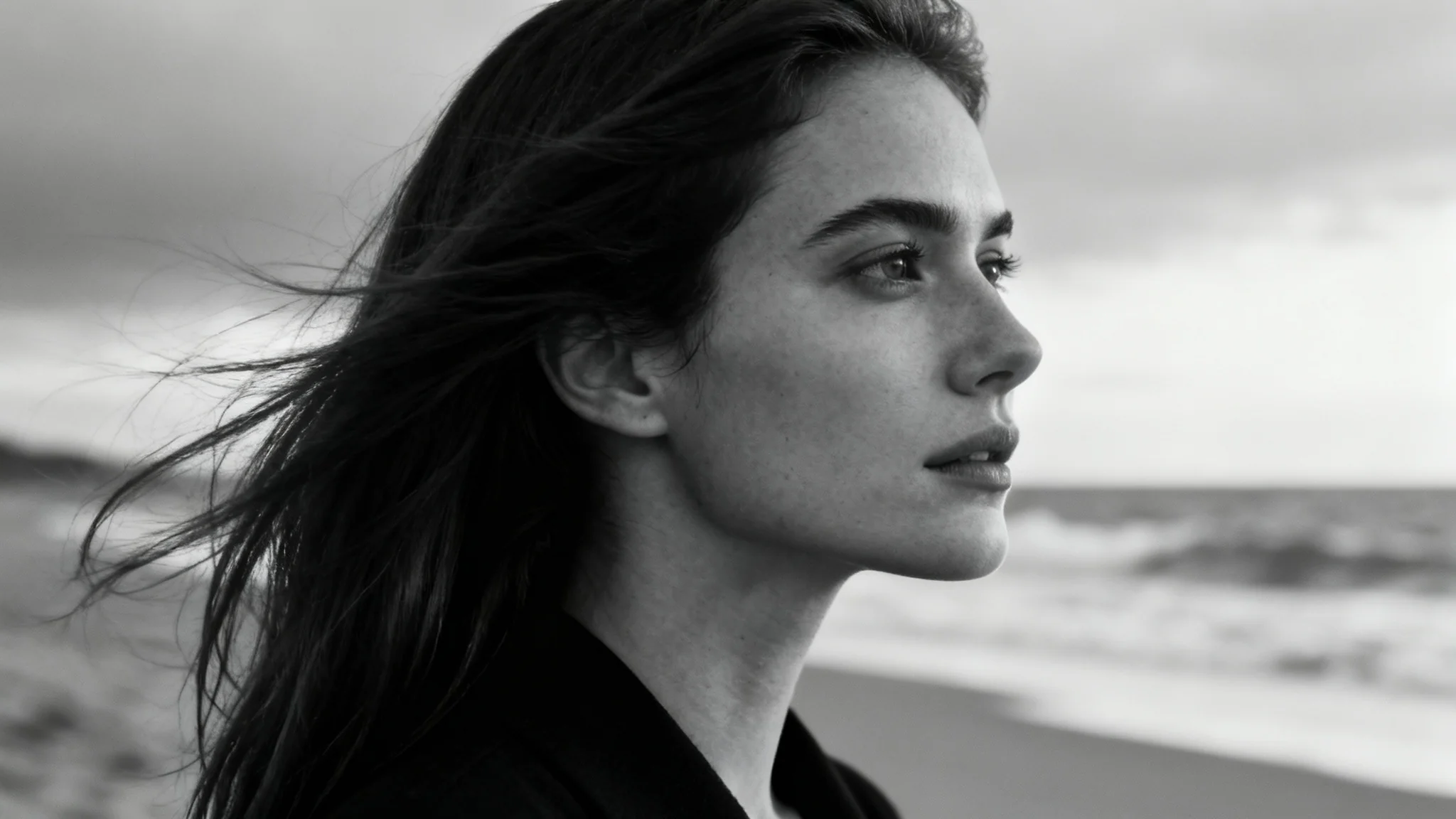 A close-up black and white portrait of a woman on a beach, her hair blowing in the wind as she looks serenely towards the ocean.