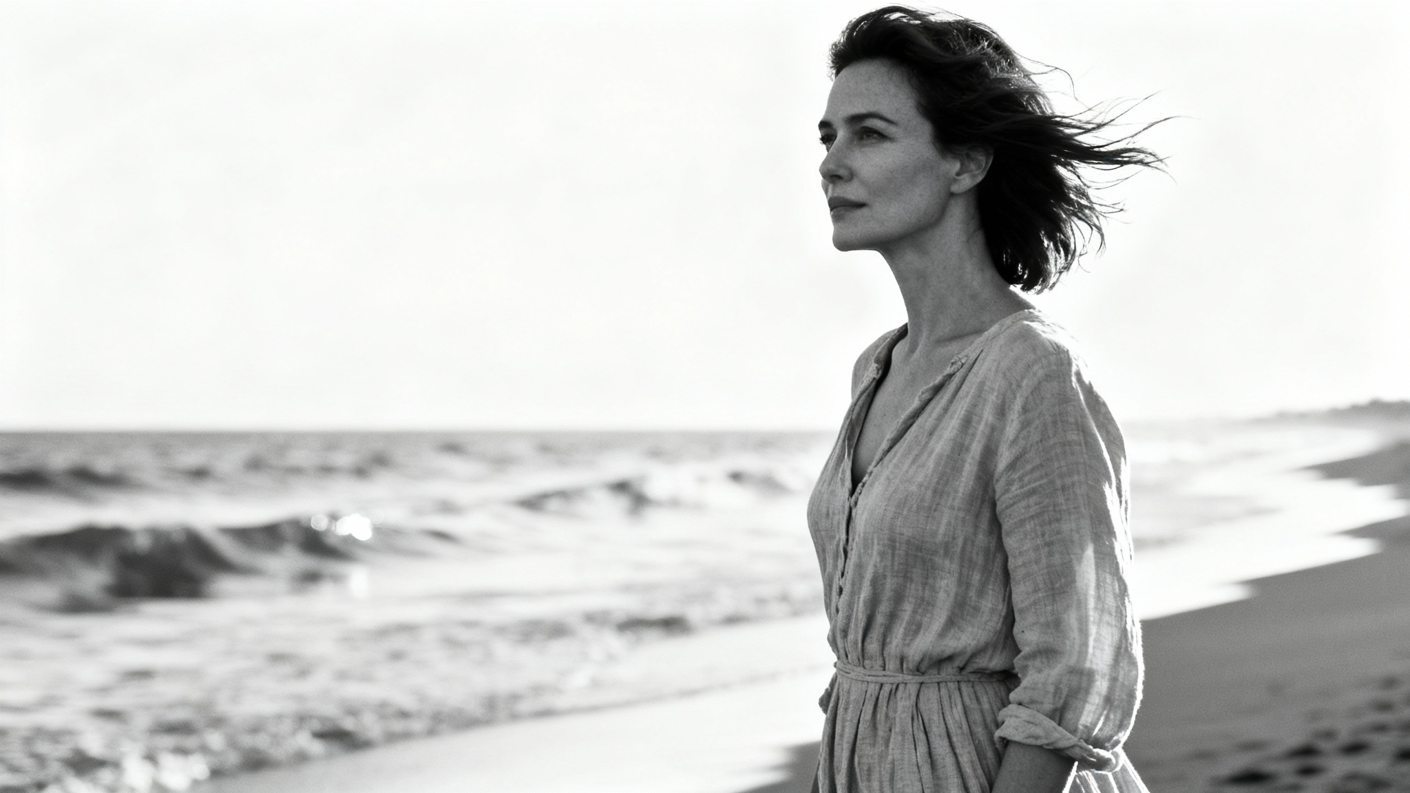 A classic black and white portrait of a woman with flowing hair standing on a serene beach, looking thoughtfully towards the ocean.