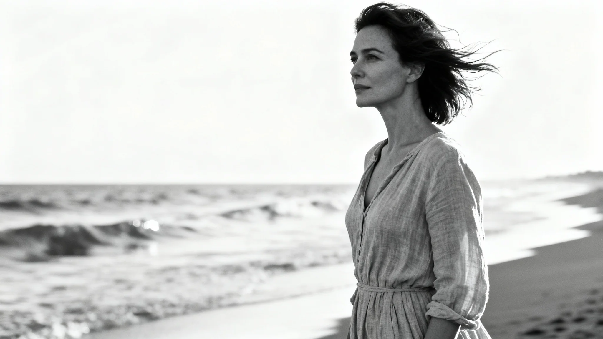 A classic black and white portrait of a woman with flowing hair standing on a serene beach, looking thoughtfully towards the ocean.