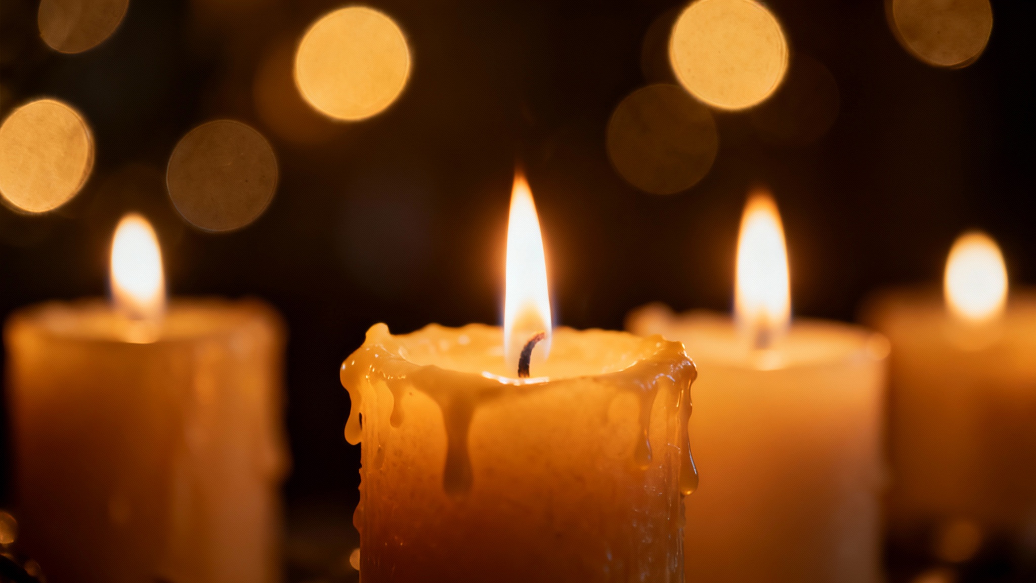 A close-up photograph of lit candles with warm, glowing flames, set against a dark background filled with soft, out-of-focus golden bokeh lights, creating a serene and cozy atmosphere.