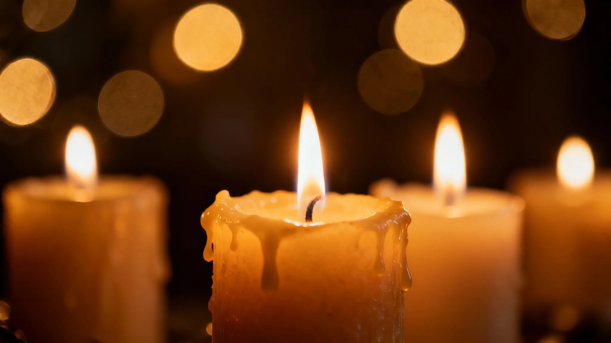A close-up photograph of lit candles with warm, glowing flames, set against a dark background filled with soft, out-of-focus golden bokeh lights, creating a serene and cozy atmosphere.