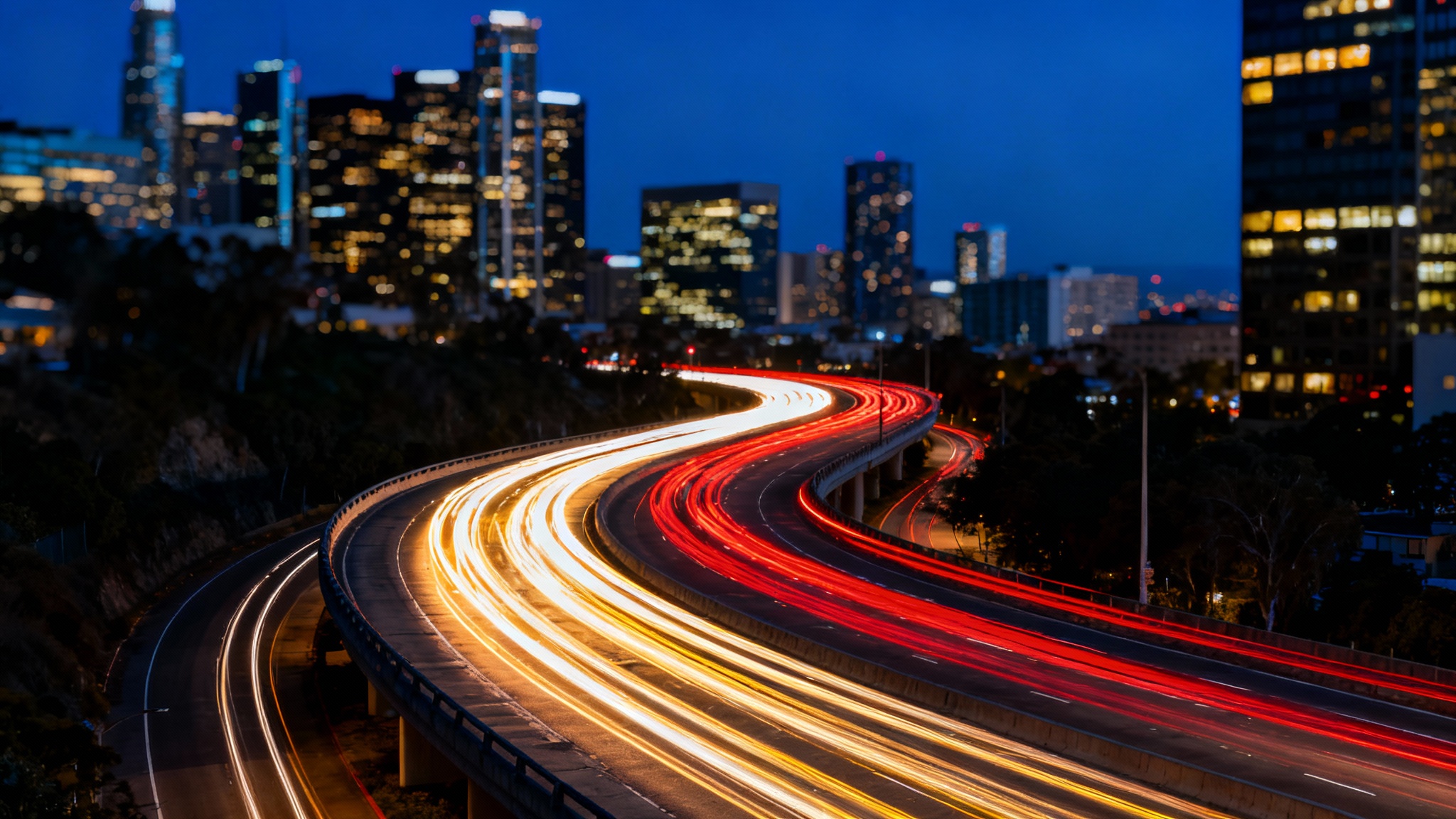 A long-exposure photograph of a city highway at night, showcasing vibrant red and white light trails from moving cars against a backdrop of illuminated skyscrapers.