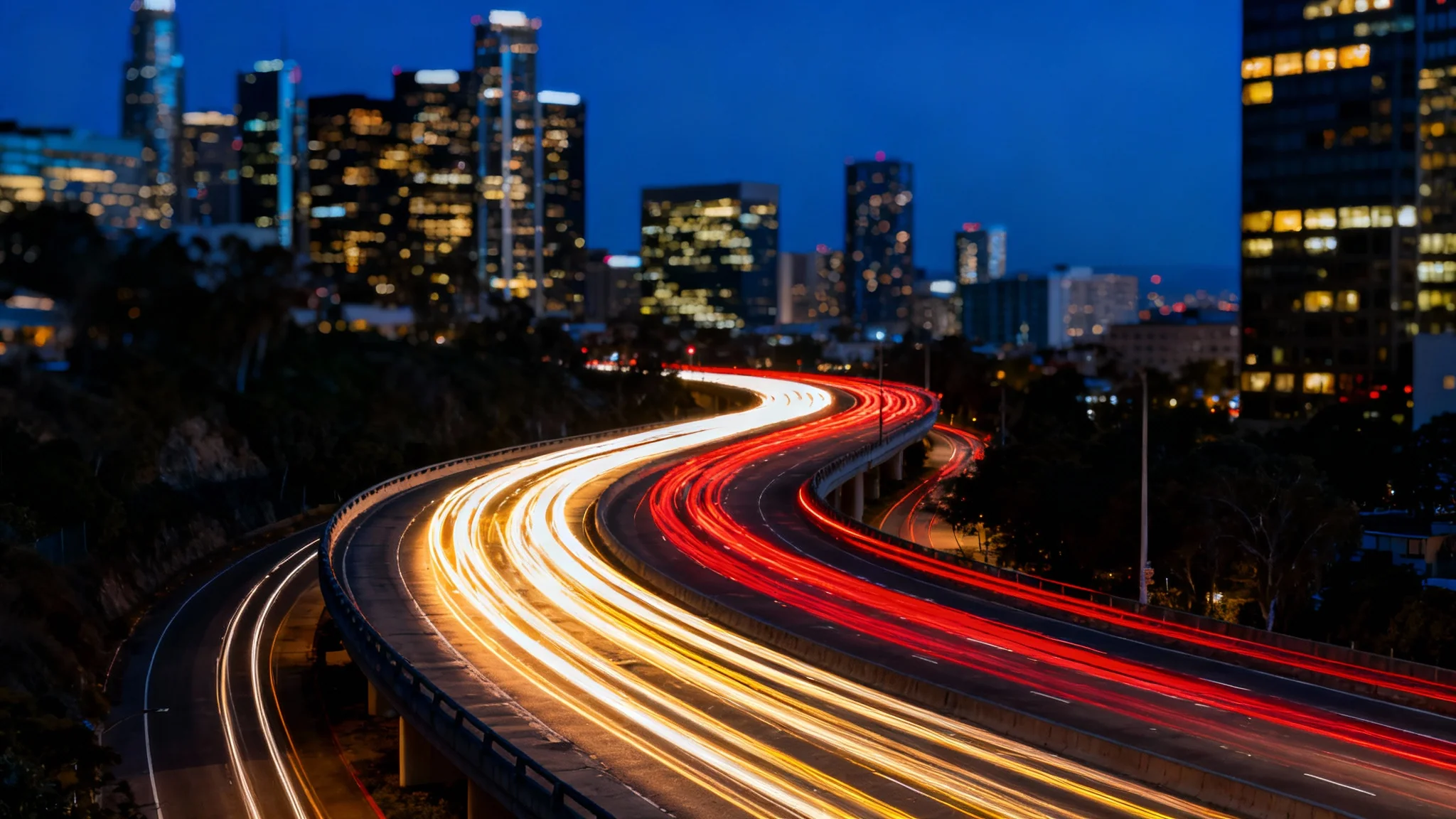 A long-exposure photograph of a city highway at night, showcasing vibrant red and white light trails from moving cars against a backdrop of illuminated skyscrapers.