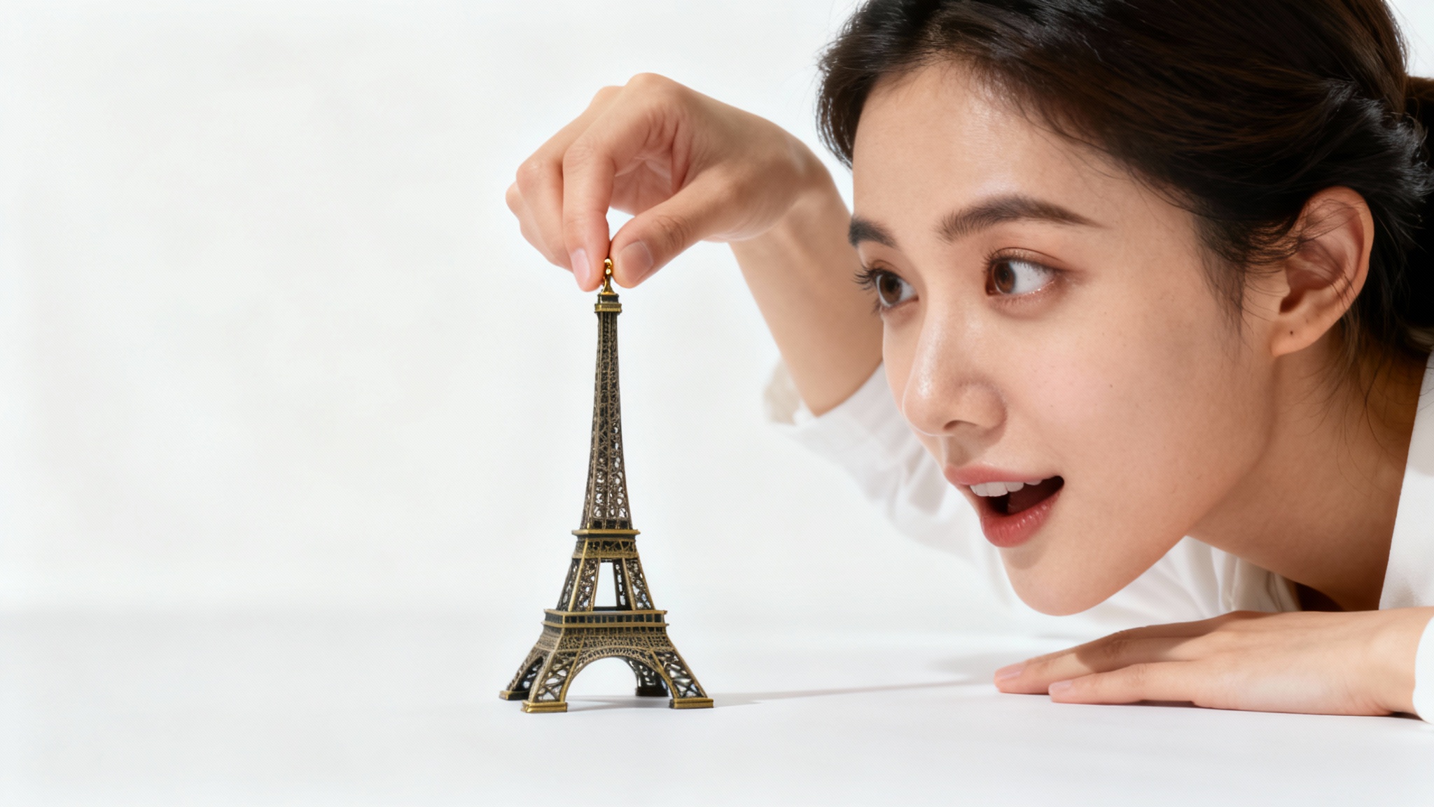 A size illusion photograph showing a woman appearing to hold a tiny version of the Eiffel Tower between her fingers against a clean white background, demonstrating forced perspective.