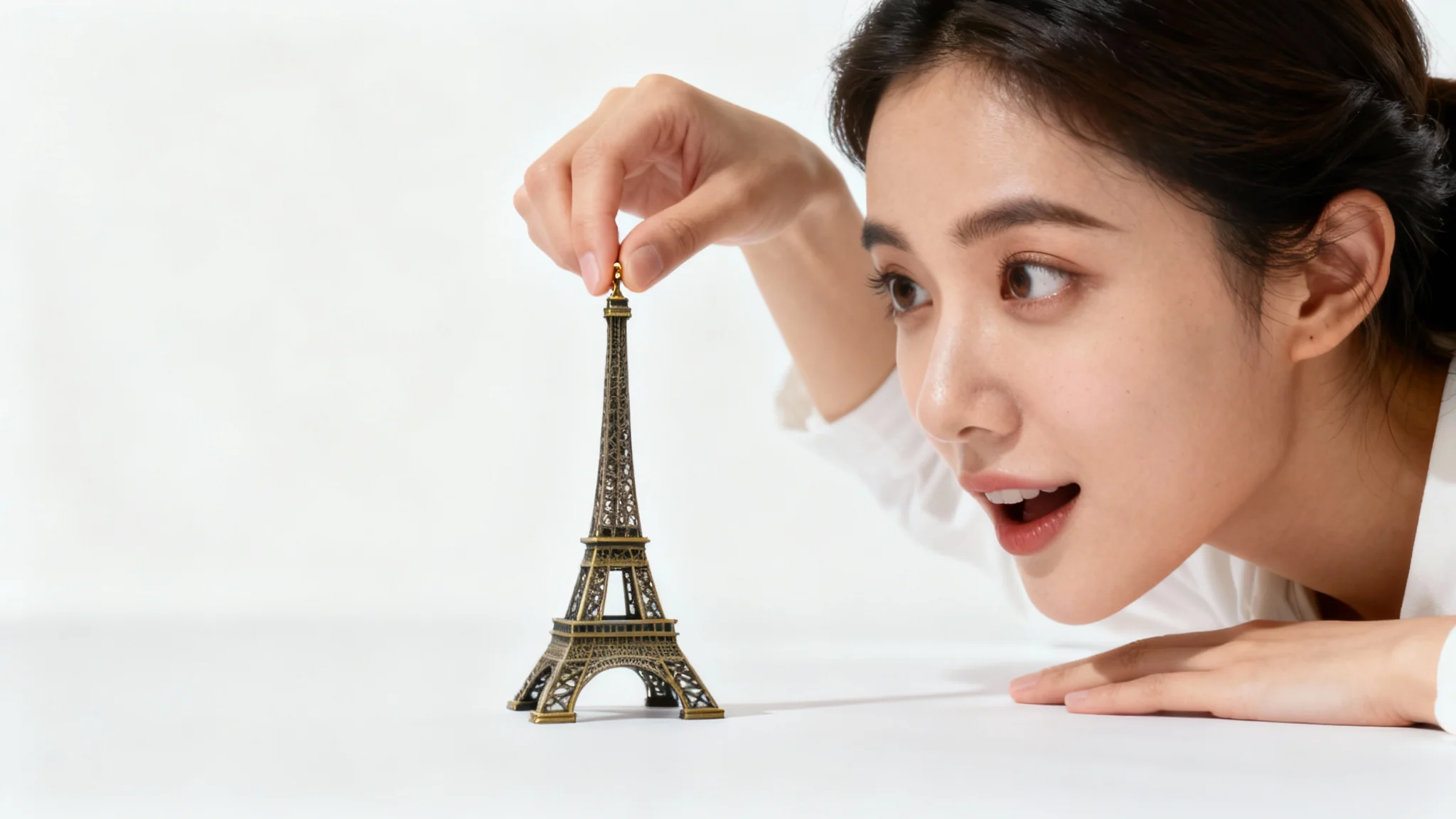 A size illusion photograph showing a woman appearing to hold a tiny version of the Eiffel Tower between her fingers against a clean white background, demonstrating forced perspective.