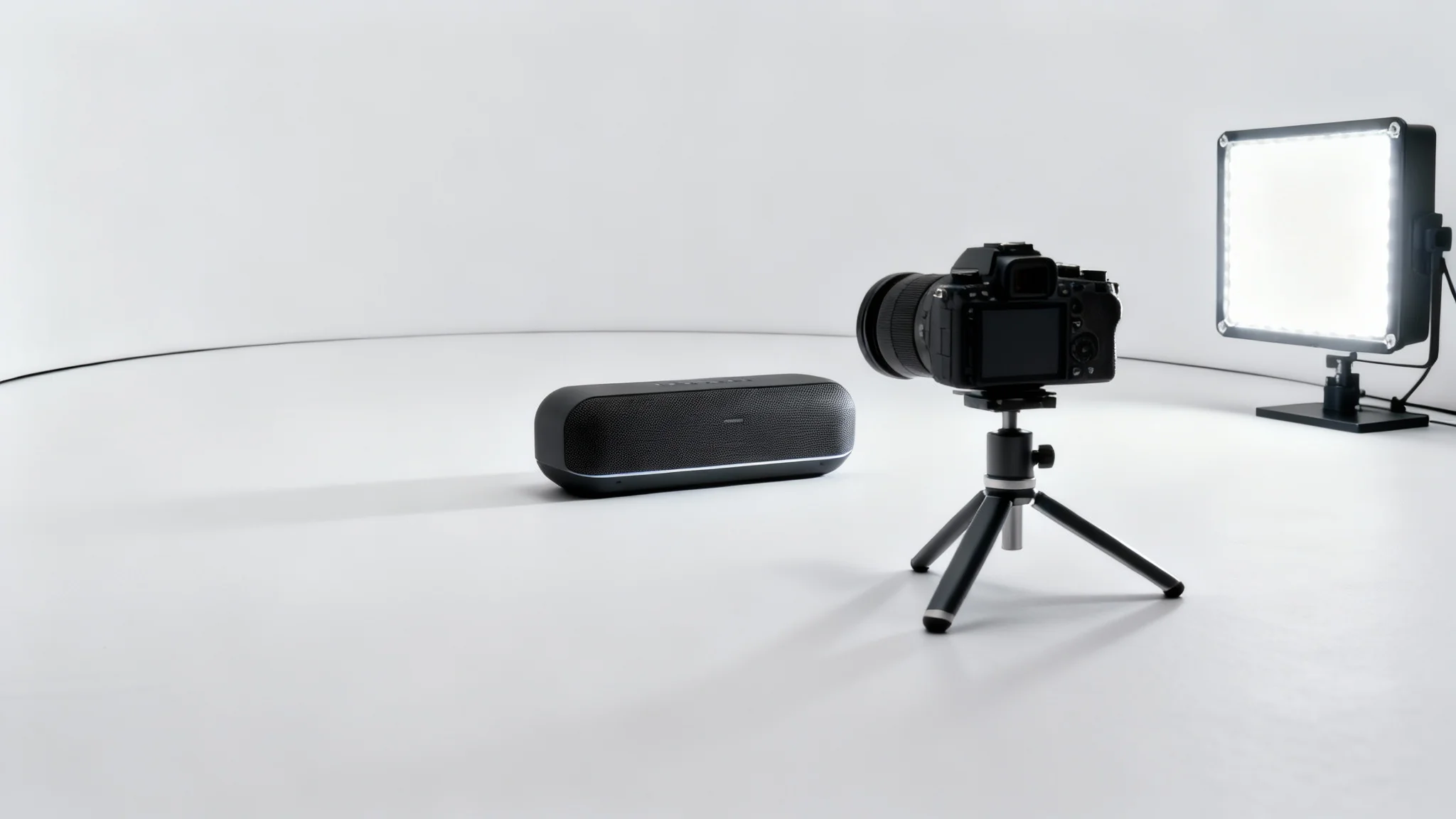 A professional product video shoot setup showing a modern wireless speaker, a camera on a tripod, and a studio light against a clean, seamless white background.