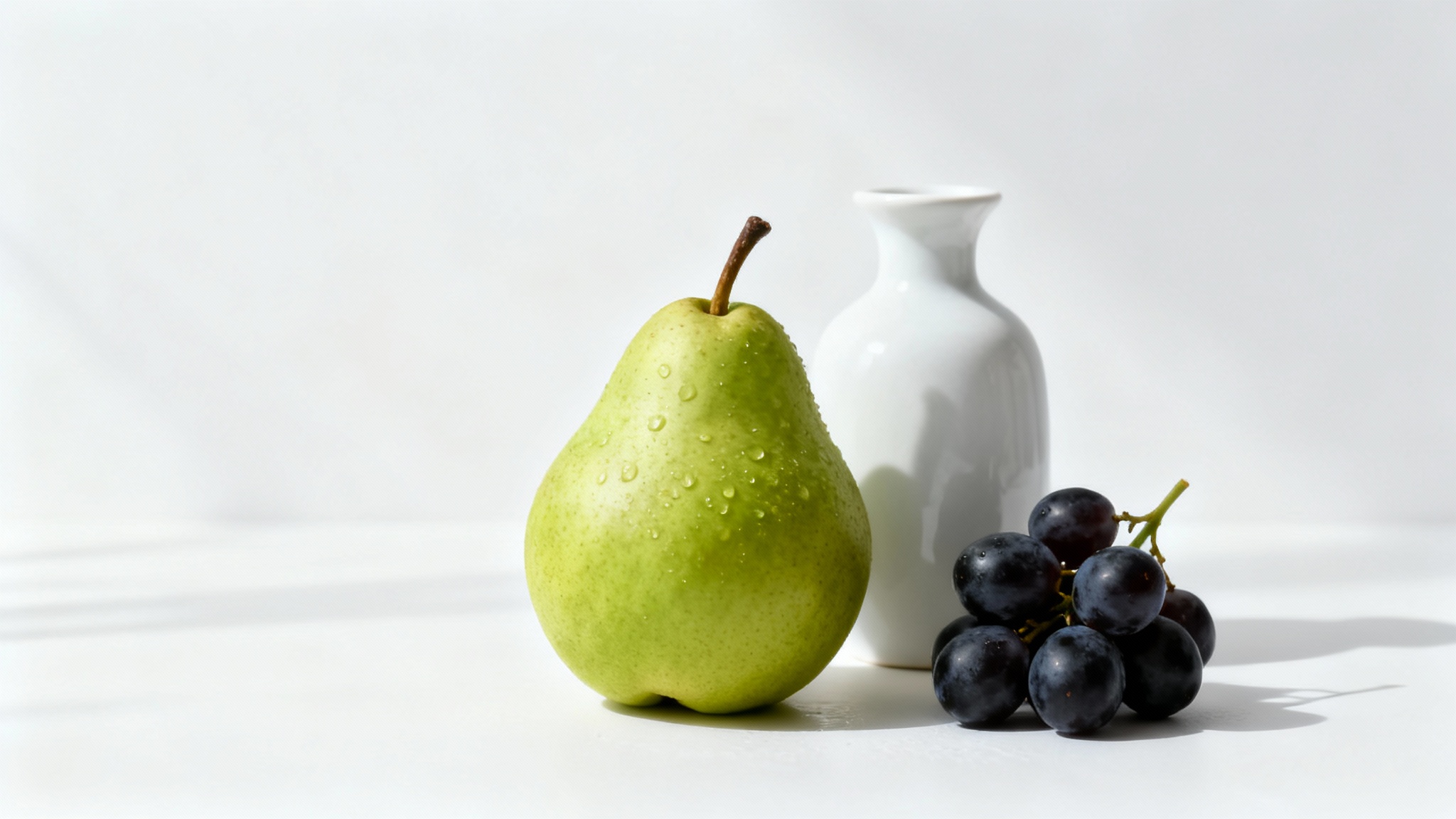 A modern still life photograph displaying a green pear, dark grapes, and a white ceramic vase arranged neatly on a surface against a clean white background.