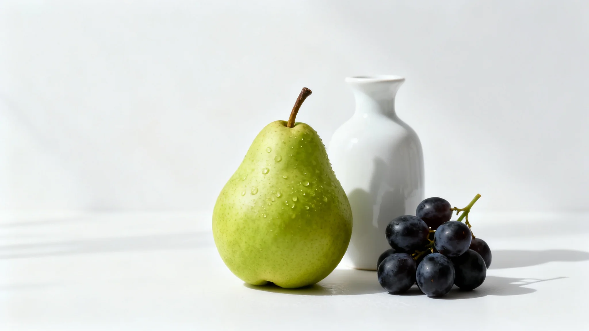 A modern still life photograph displaying a green pear, dark grapes, and a white ceramic vase arranged neatly on a surface against a clean white background.