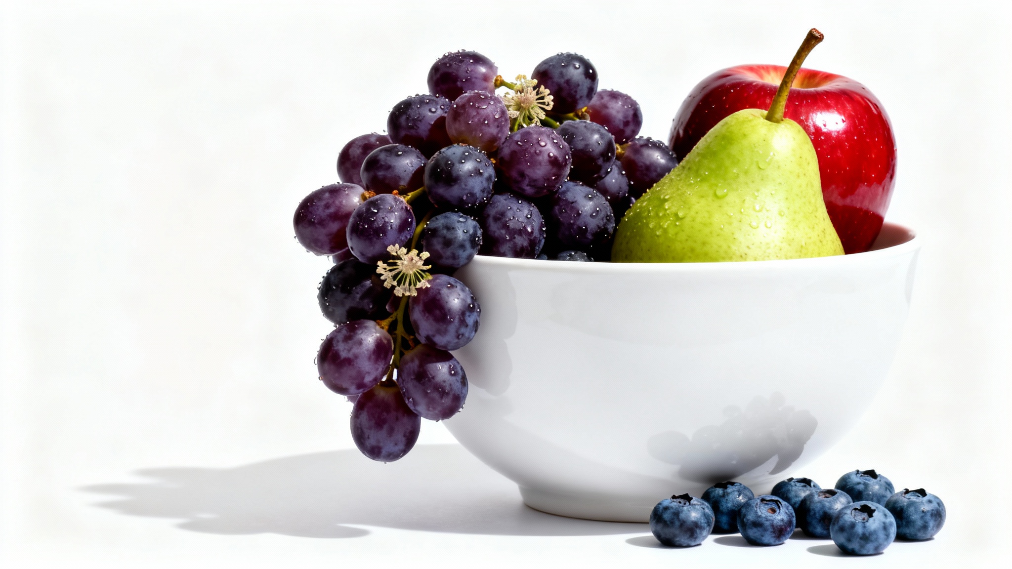 A modern still life photo of a white ceramic bowl filled with fresh grapes, a red apple, and a green pear, shot in a studio setting against a pure white background.
