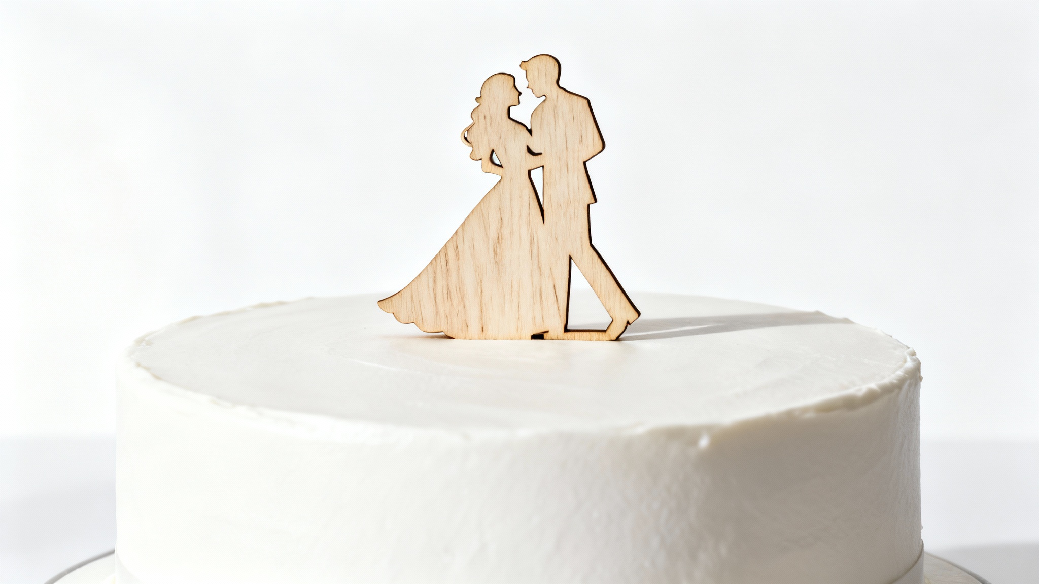A close-up product shot of a minimalist white wedding cake featuring a custom-made natural wood topper carved into the silhouette of a dancing couple, set against a plain white background.