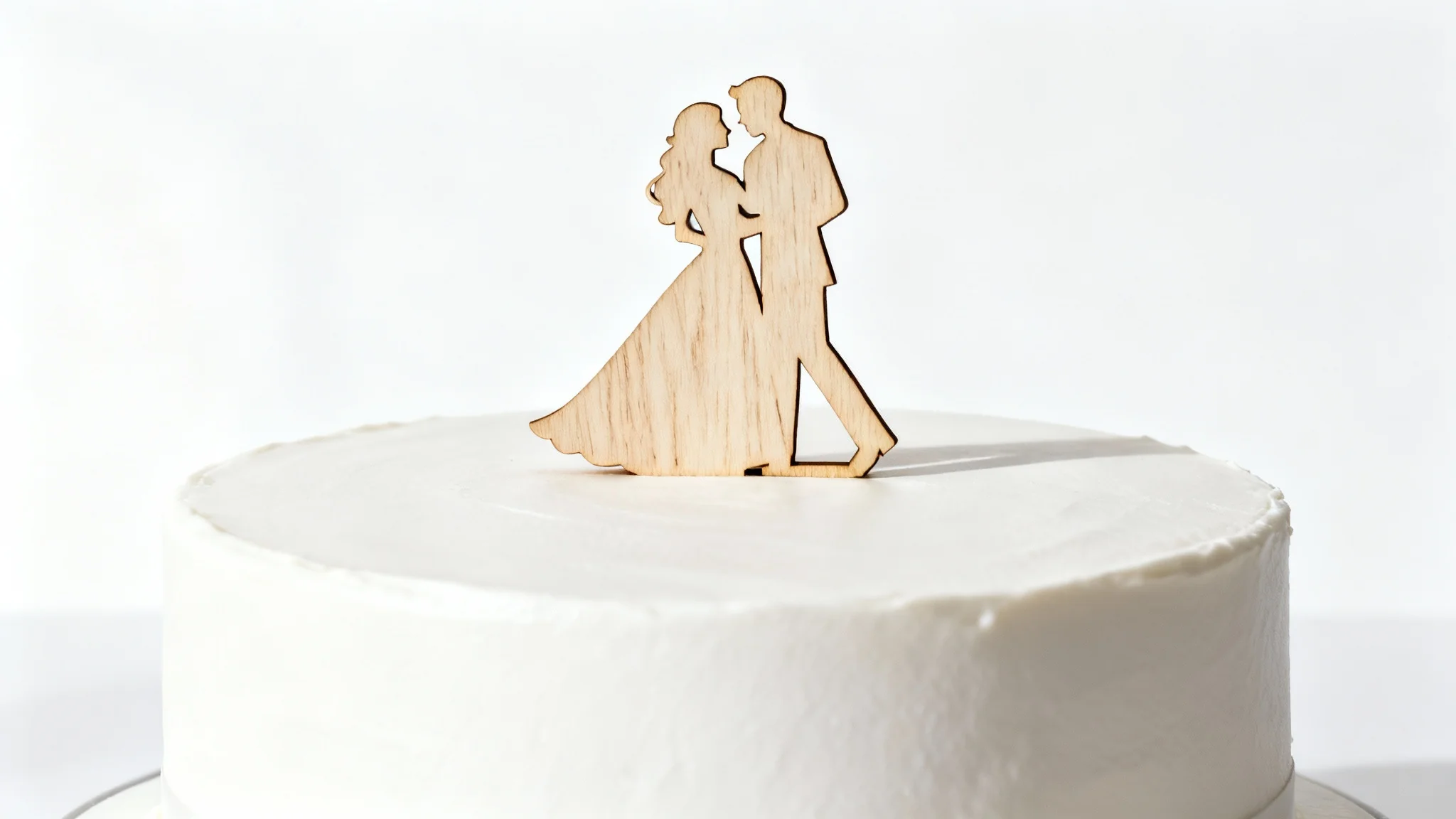 A close-up product shot of a minimalist white wedding cake featuring a custom-made natural wood topper carved into the silhouette of a dancing couple, set against a plain white background.