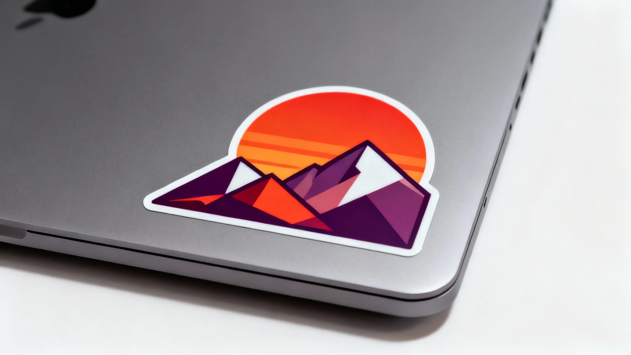 A modern space-gray laptop on a white background, featuring a colorful, die-cut sticker of a geometric mountain sunset on its lid.