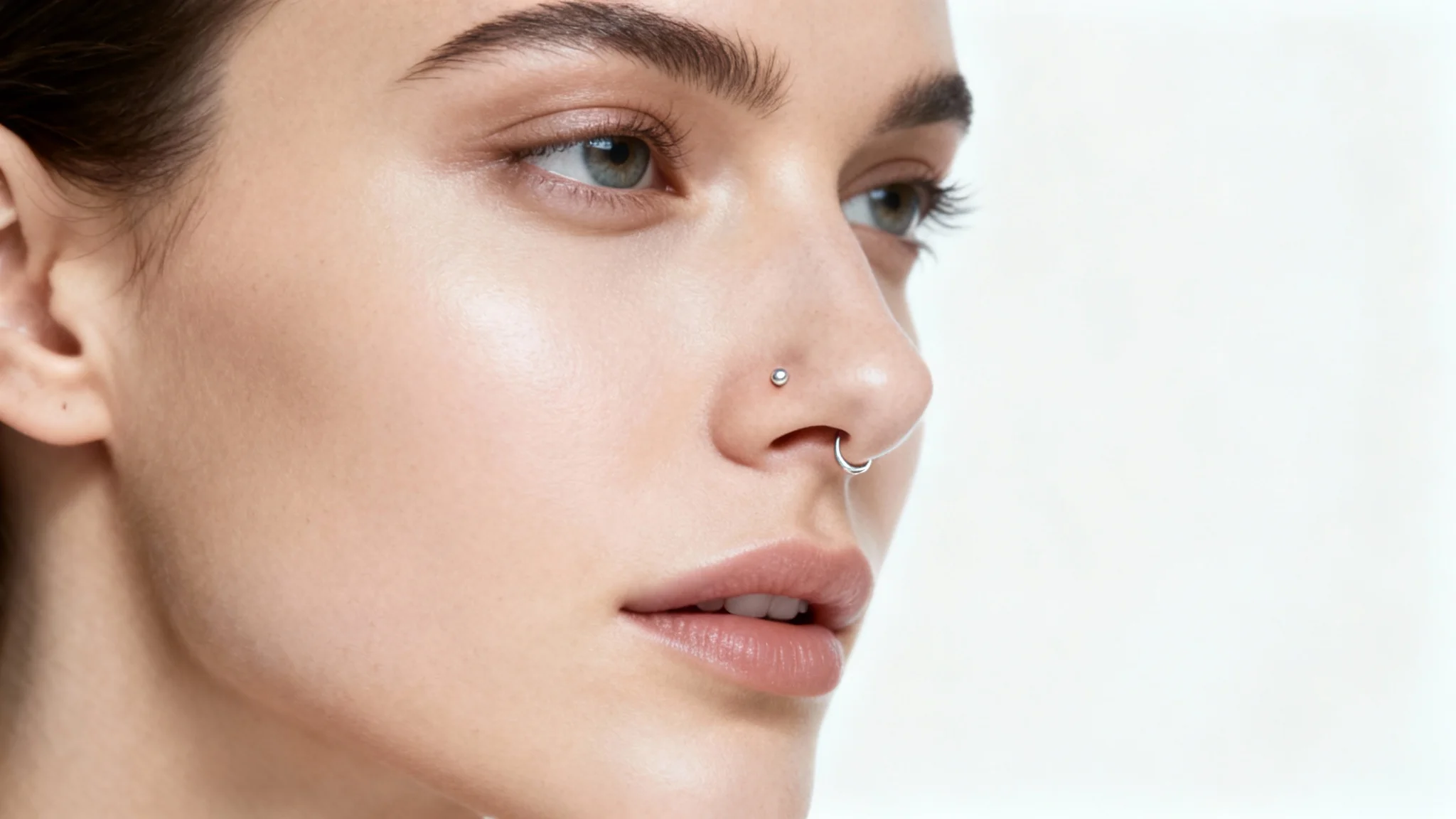 A close-up studio photograph of a person's face in profile, highlighting a delicate silver stud piercing in their nostril against a clean white background.