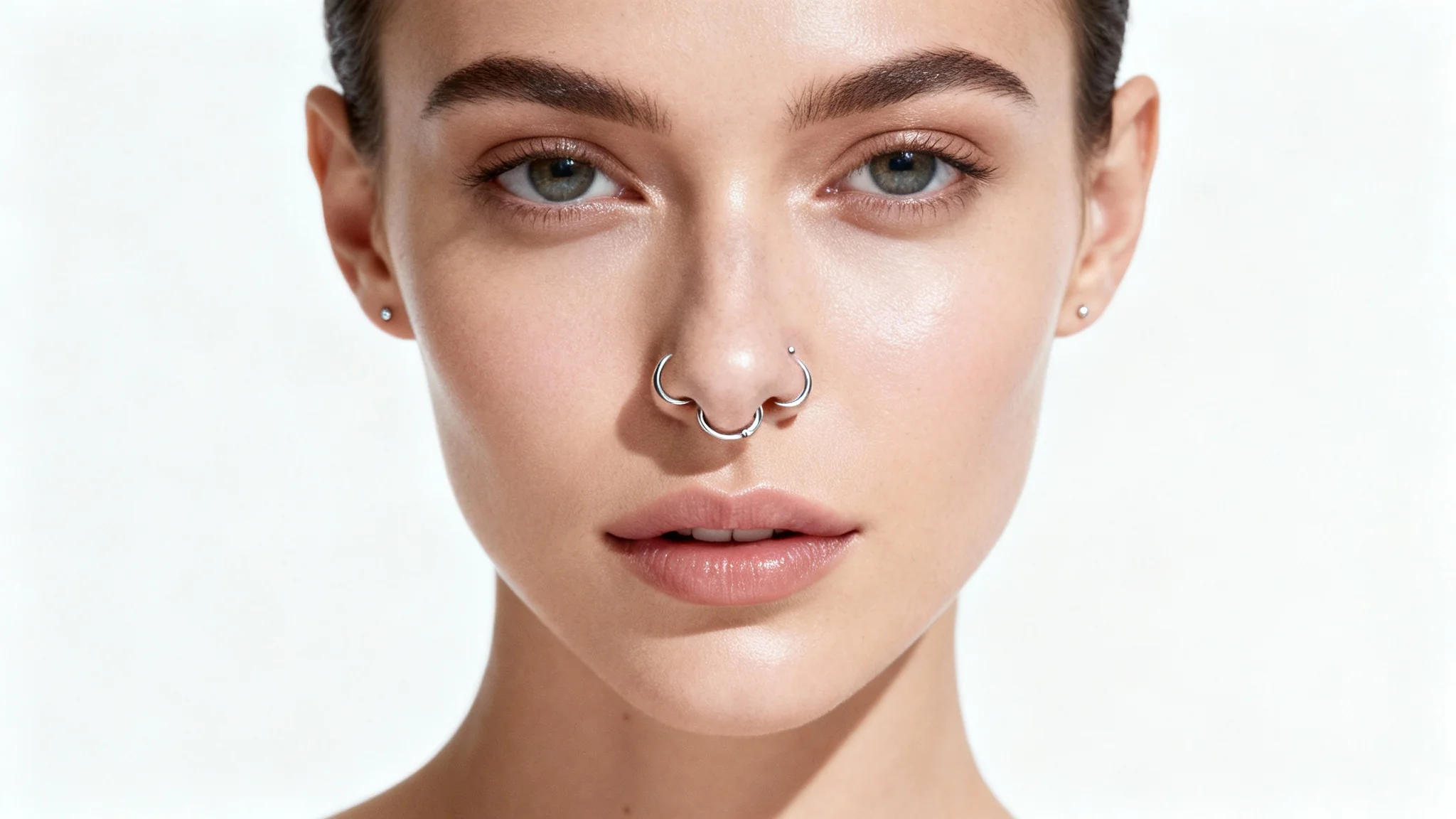 A close-up, high-fashion portrait of a person's face against a white background, showcasing a delicate silver septum ring and a matching nostril stud.