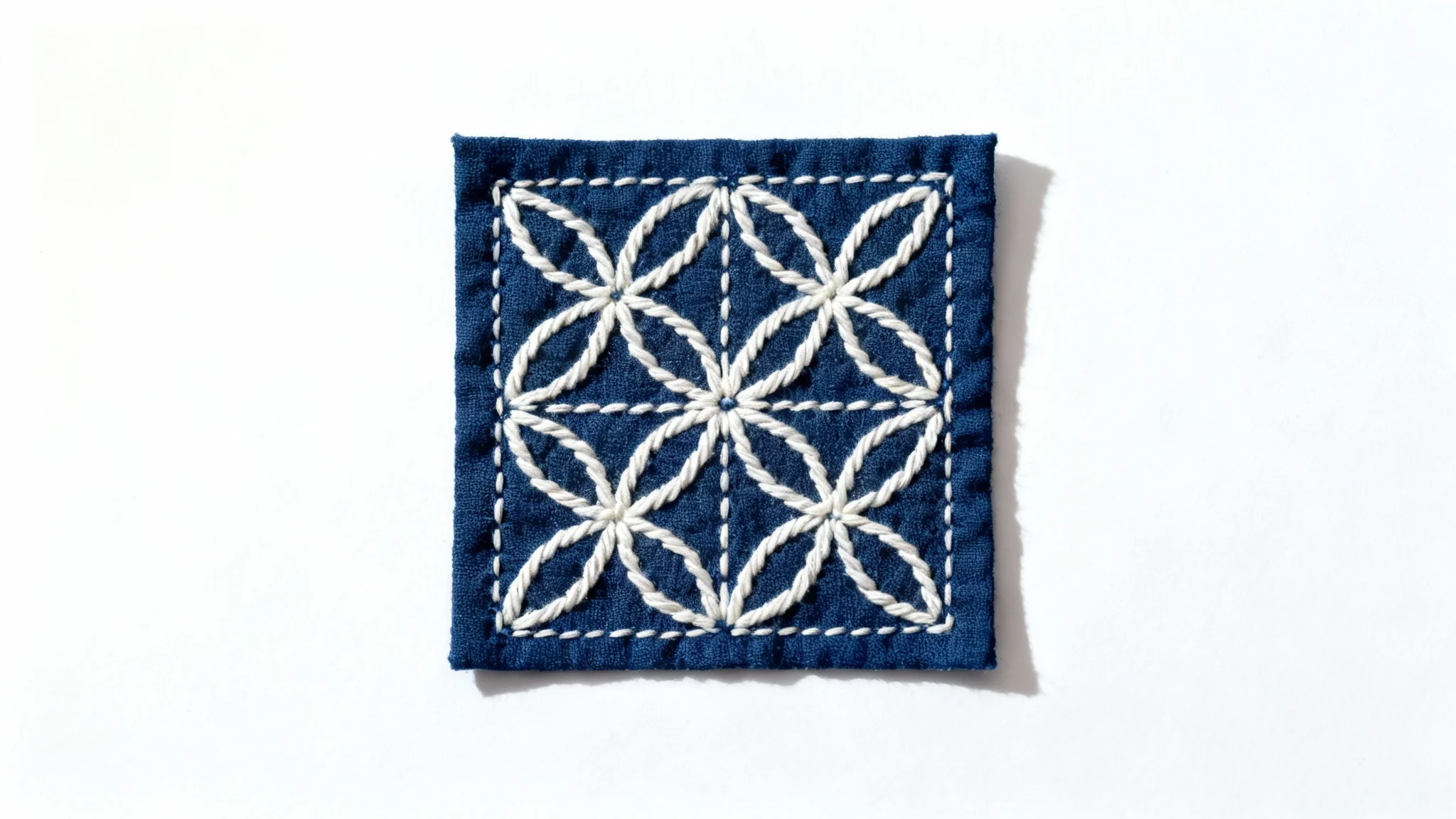 A close-up, top-down view of a piece of indigo-dyed fabric featuring a traditional Japanese Sashiko pattern of interlocking hemp leaves stitched in thick white thread, set against a plain white background.