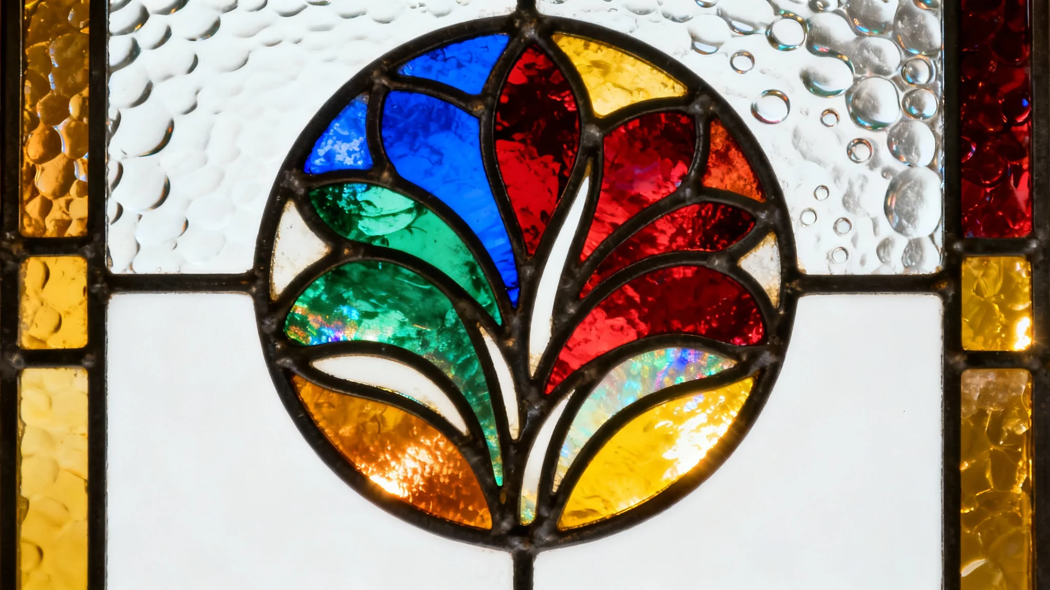 A close-up, photorealistic image of a circular Art Nouveau-style stained glass panel featuring a floral design in vibrant jewel tones, backlit against a plain white background.