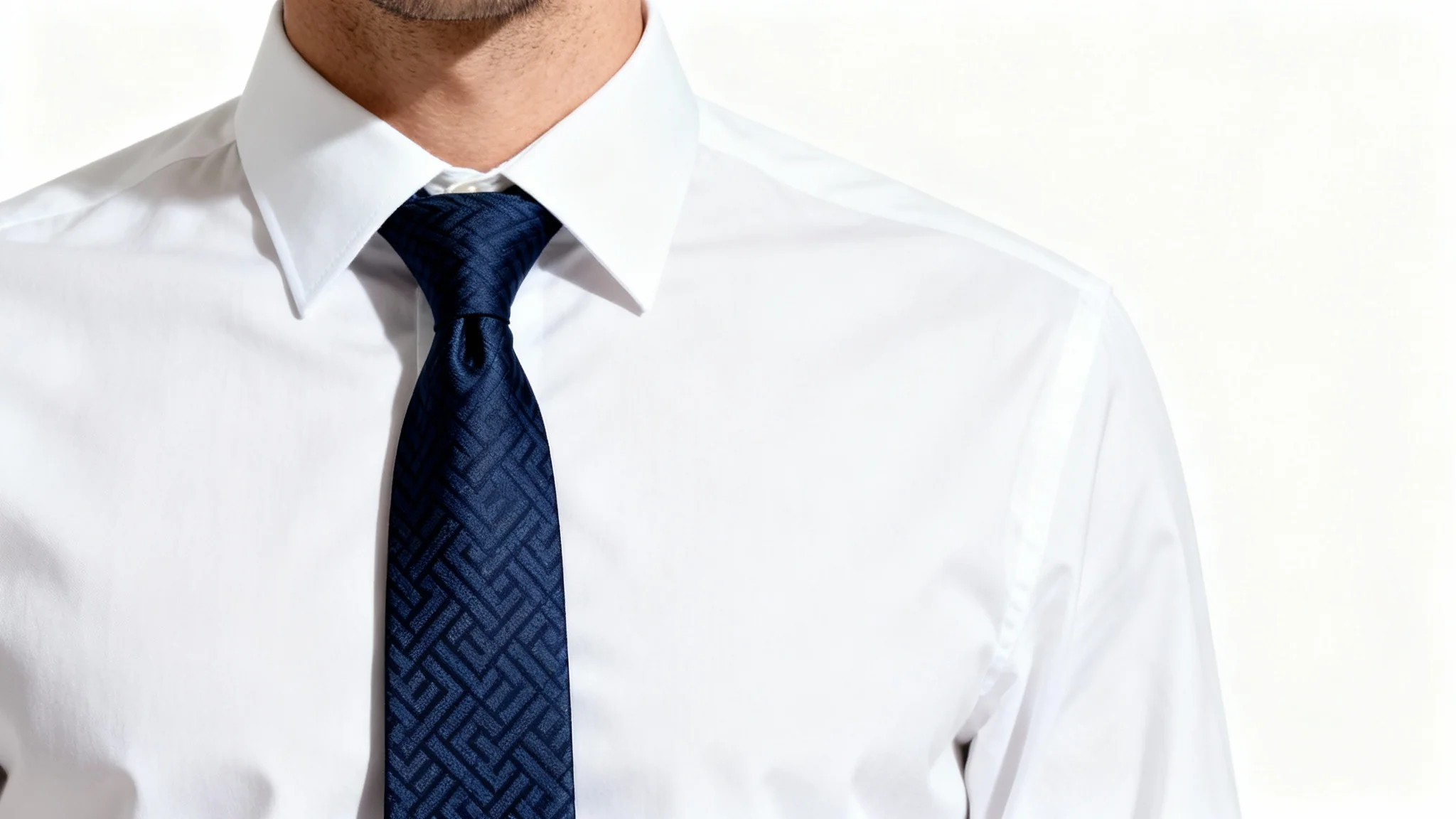 A close-up studio photograph of a man's chest, showing him wearing a crisp white shirt and a perfectly knotted, elegant navy blue silk tie against a plain white background.