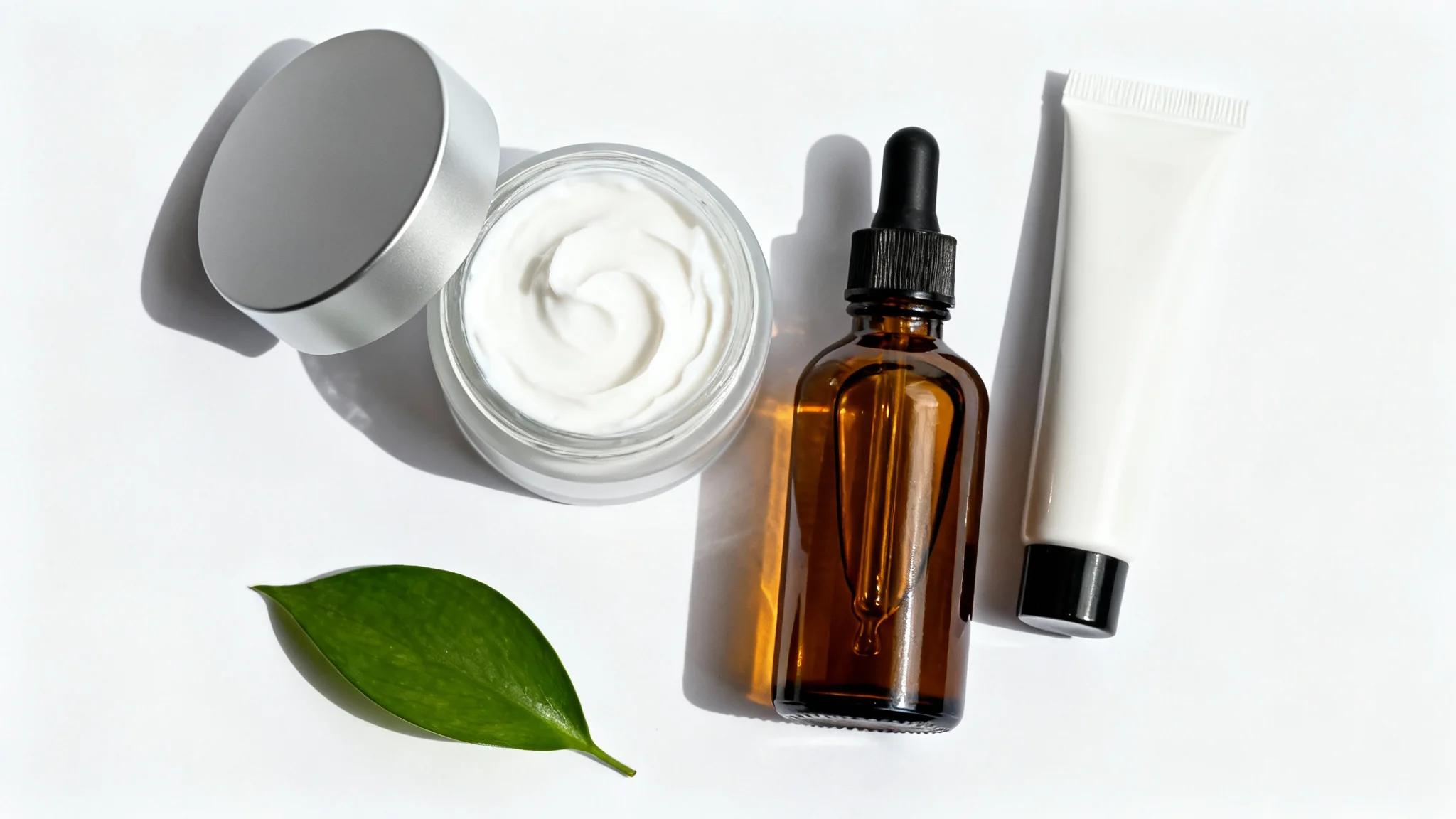 A minimalist top-down flat lay of cosmetic products, including a jar of cream and a serum bottle, arranged on a clean white background with a single green leaf.