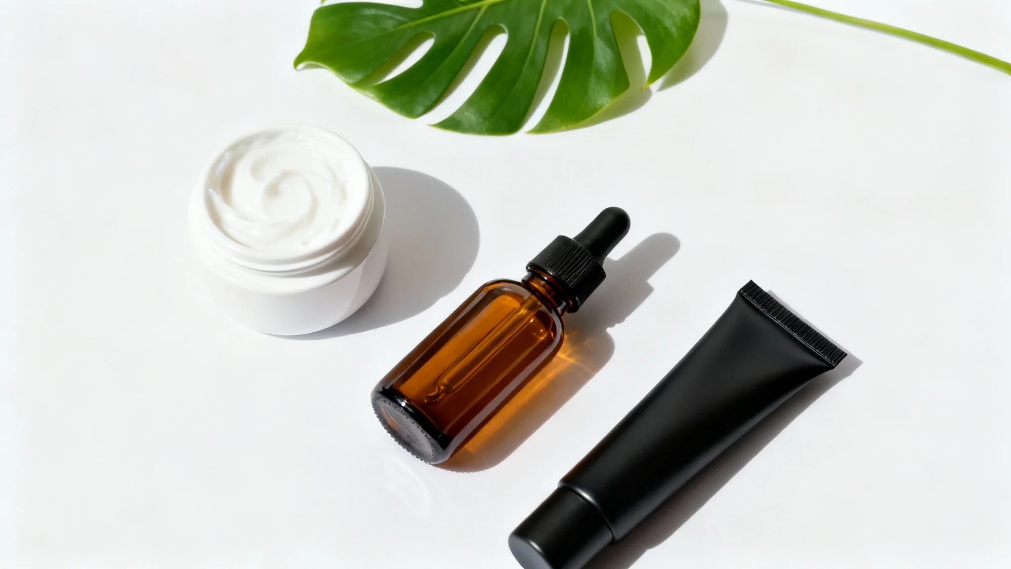 A sophisticated top-down flat lay of unbranded cosmetic products, including a cream jar and a serum bottle, arranged neatly on a clean white background with a single green leaf.