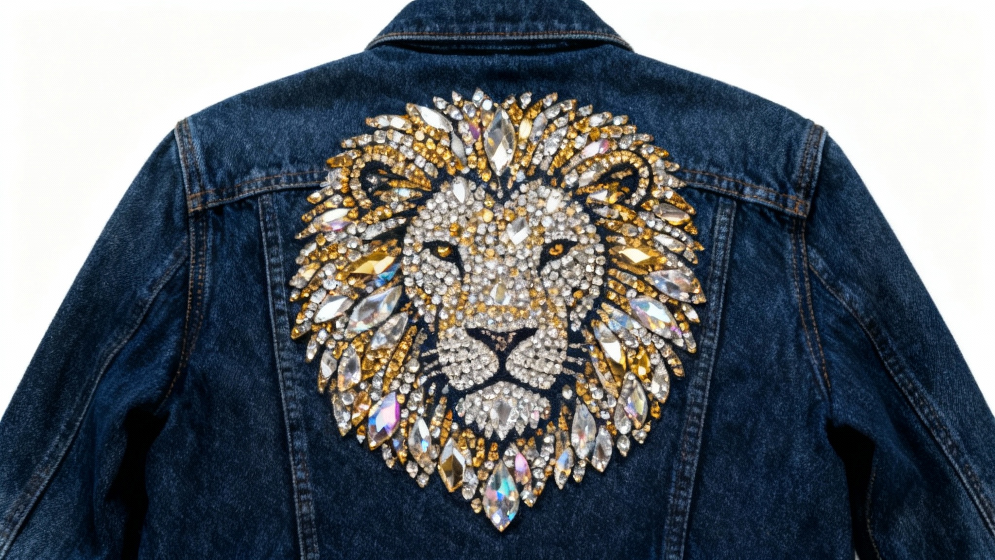 A close-up of a dark blue denim jacket with a highly detailed, sparkling rhinestone pattern of a lion's head on the back, laid flat on a white background.