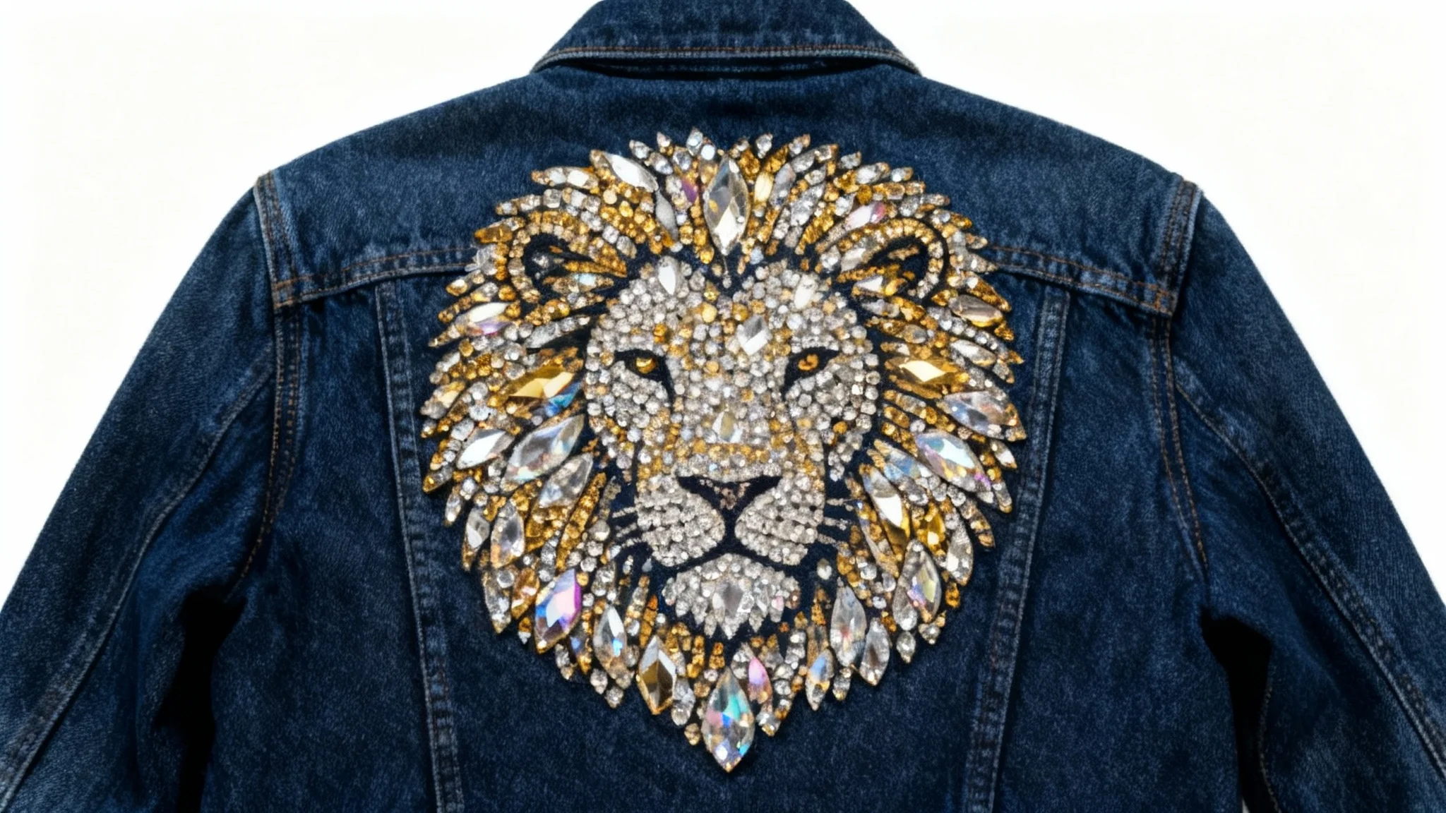 A close-up of a dark blue denim jacket with a highly detailed, sparkling rhinestone pattern of a lion's head on the back, laid flat on a white background.