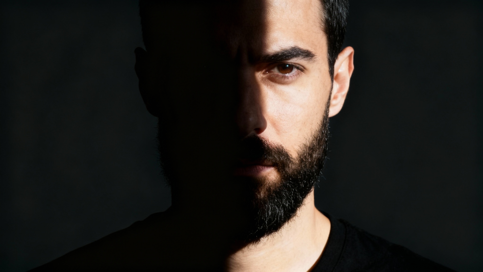 A dramatic 16:9 studio portrait of a man demonstrating a perfect split lighting effect, where one side of his face is brightly lit and the other is in deep shadow, creating a striking contrast.