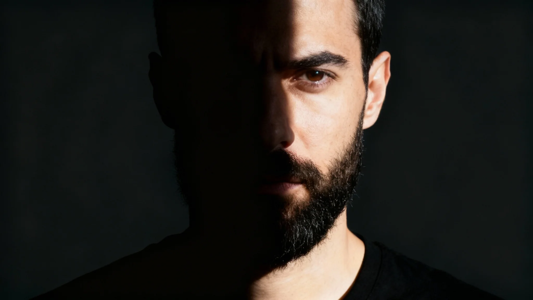 A dramatic 16:9 studio portrait of a man demonstrating a perfect split lighting effect, where one side of his face is brightly lit and the other is in deep shadow, creating a striking contrast.
