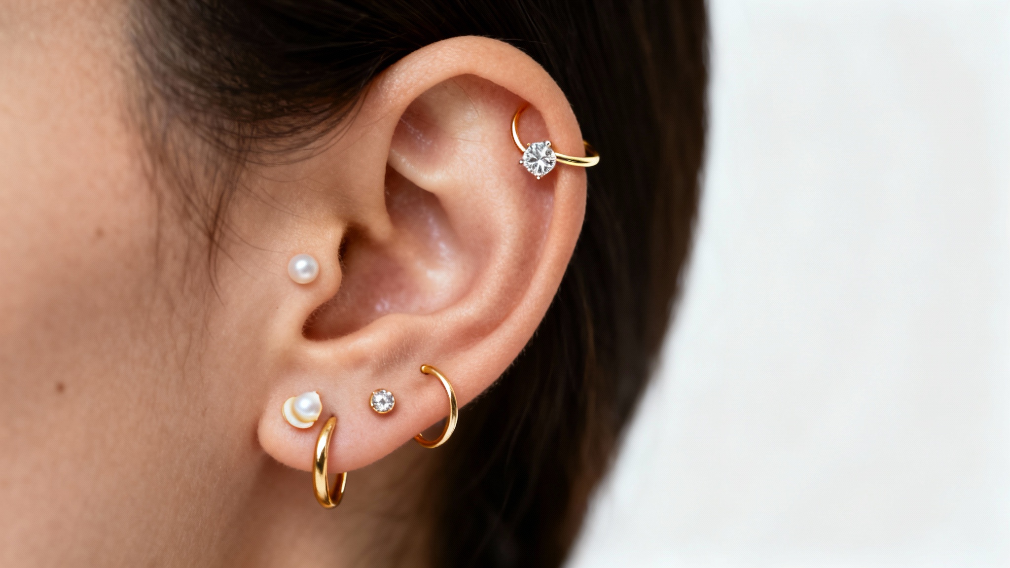 A close-up studio photograph of an ear elegantly decorated with multiple piercings, including a gold hoop, diamond stud, and a pearl, shown against a plain white background as a mockup.