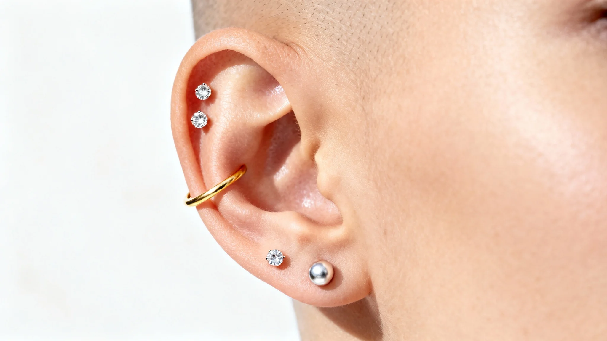 A close-up, high-fashion photograph of a human ear showcasing multiple elegant piercings, including diamond studs and a gold hoop, set against a stark white background.
