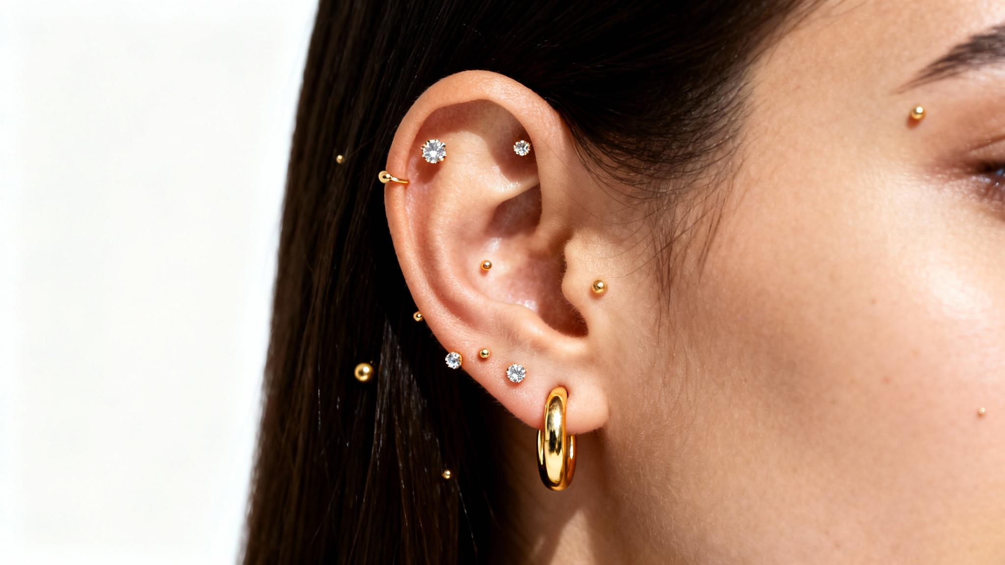 A close-up image of a person's ear with multiple piercings, featuring a stylish arrangement of small gold earrings against a clean white background.