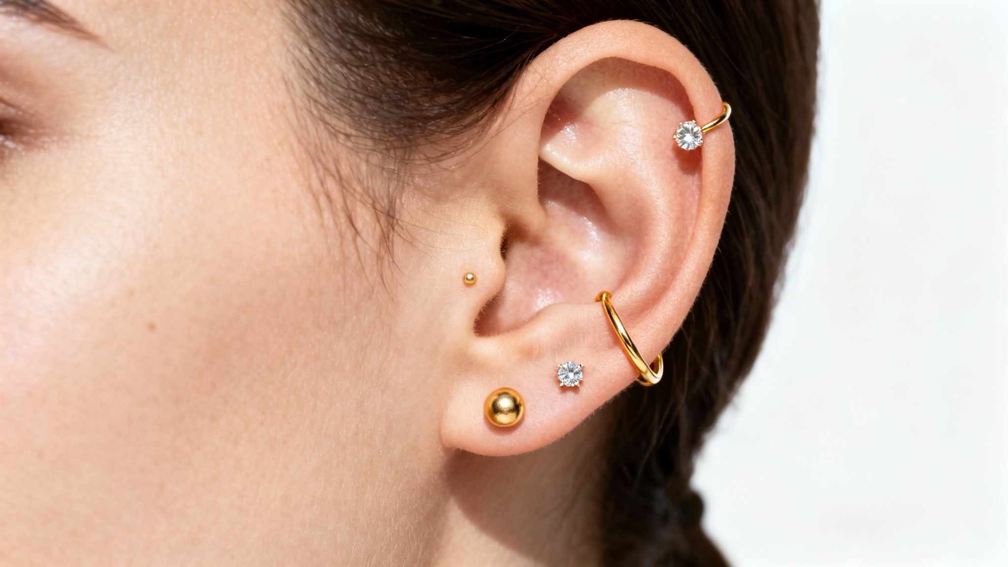 A hyper-realistic close-up of a human ear showcasing a mockup of three piercings: a small gold hoop in the lobe, a diamond stud in the helix, and a gold stud in the conch, all set against a clean white background.