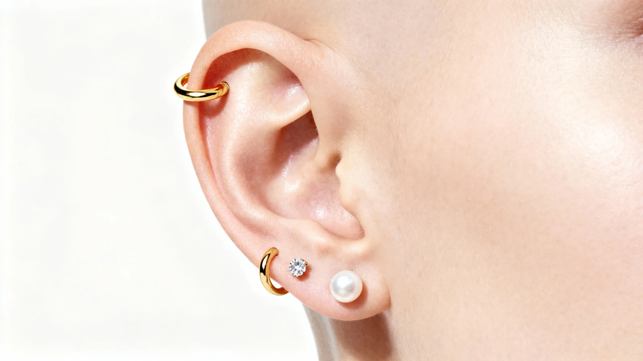 A highly detailed close-up of a human ear featuring mockups of various piercings: a gold hoop in the helix, a diamond stud in the tragus, and a pearl in the lobe, all set against a stark white background.