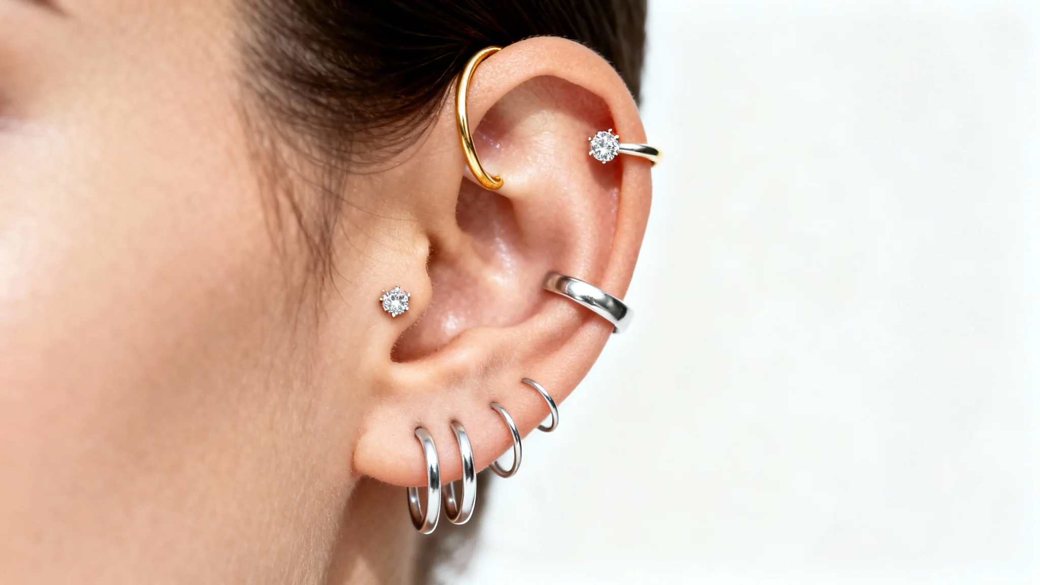 A close-up photo of a person's ear adorned with various stylish piercings, including gold hoops, a silver cuff, and a diamond stud, set against a clean white background.
