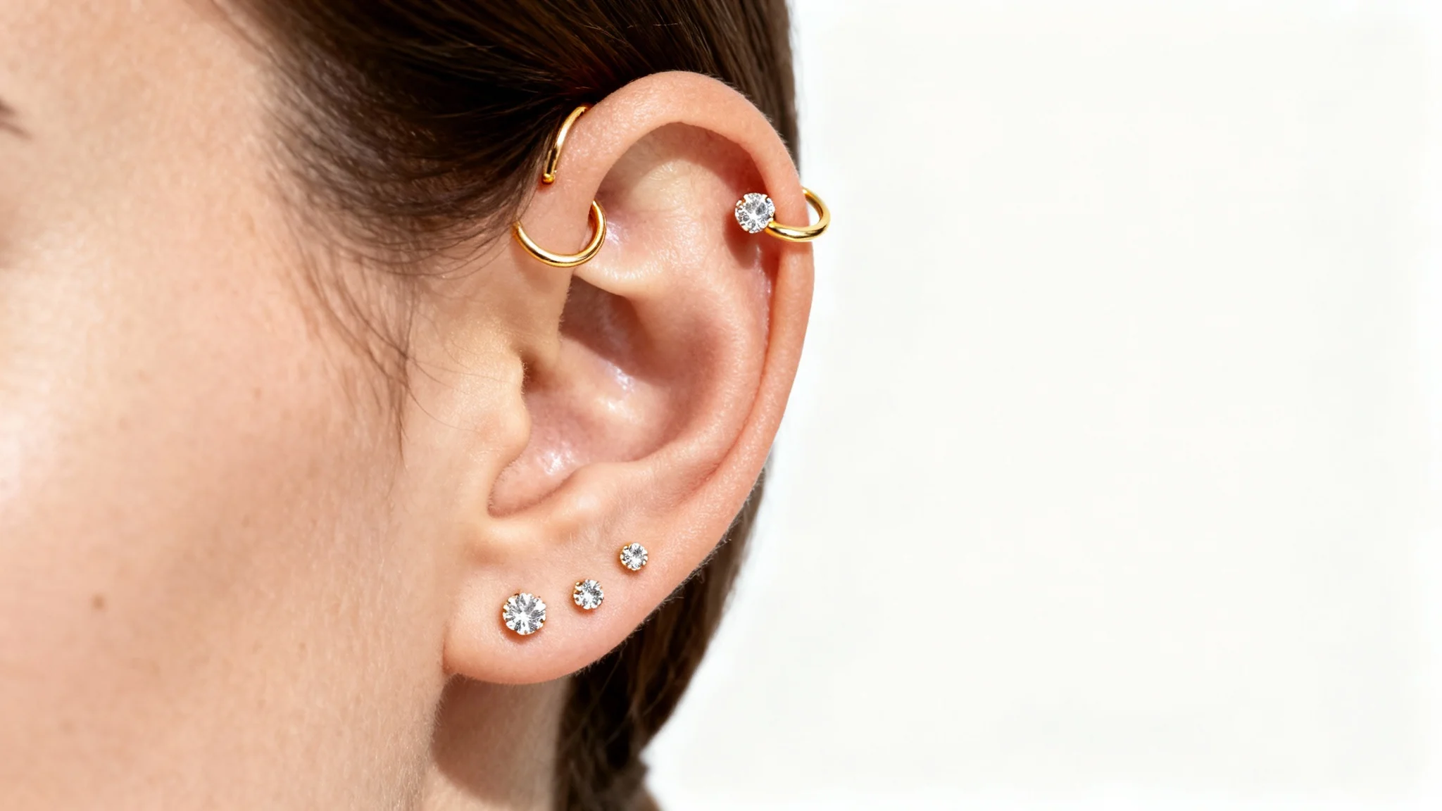 A close-up, high-detail photograph of a woman's ear adorned with multiple delicate gold and diamond piercings, shot against a clean white background.