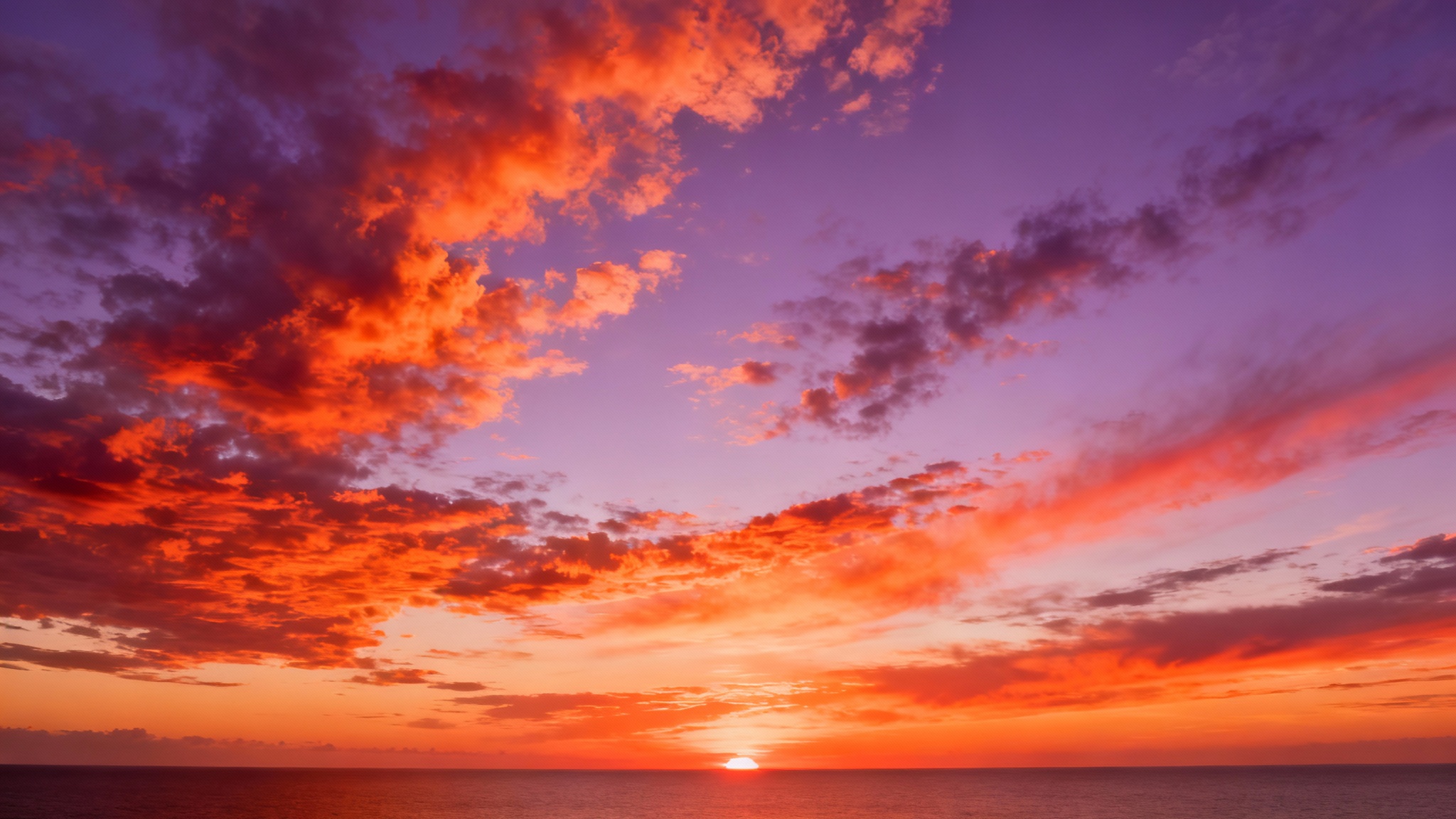A vibrant 360-degree panoramic image of a sunset sky, shown as a flat equirectangular projection. The sky is filled with clouds colored in shades of orange, purple, and red by the setting sun.